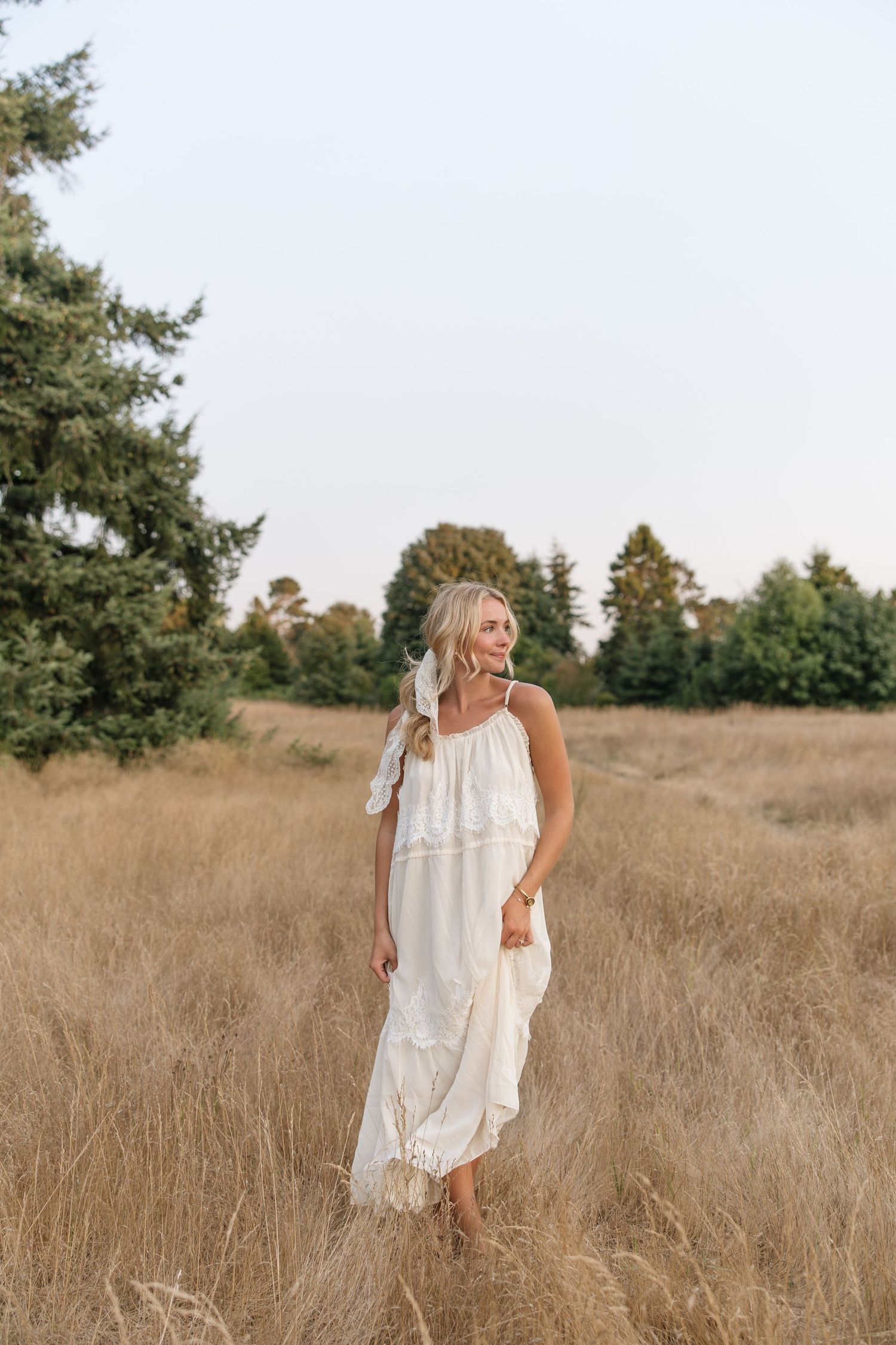 Woman in white boho maxi dress standing in golden field with evergreen trees in background.