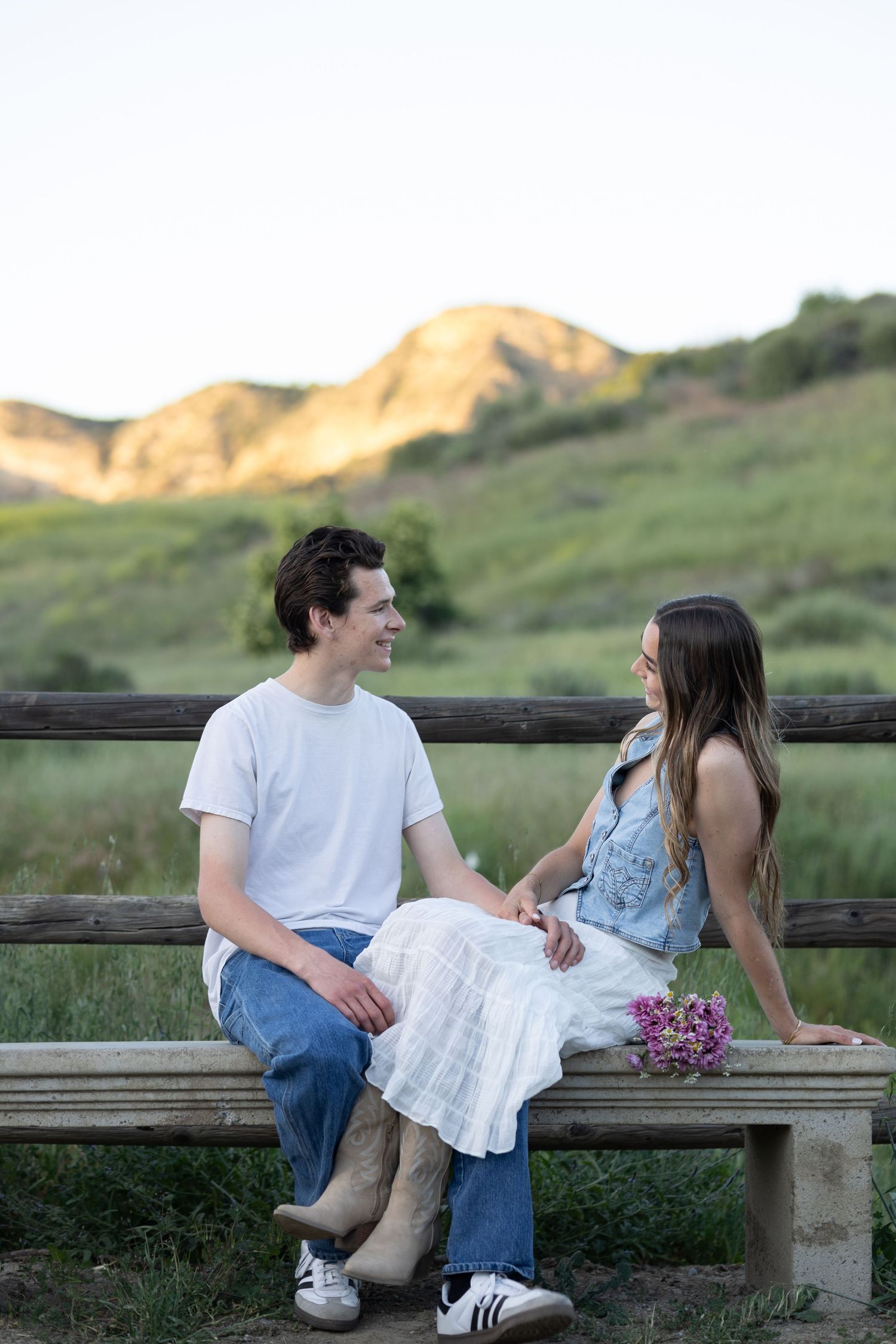 Couple sitting on wooden bench against mountain backdrop at golden hour.