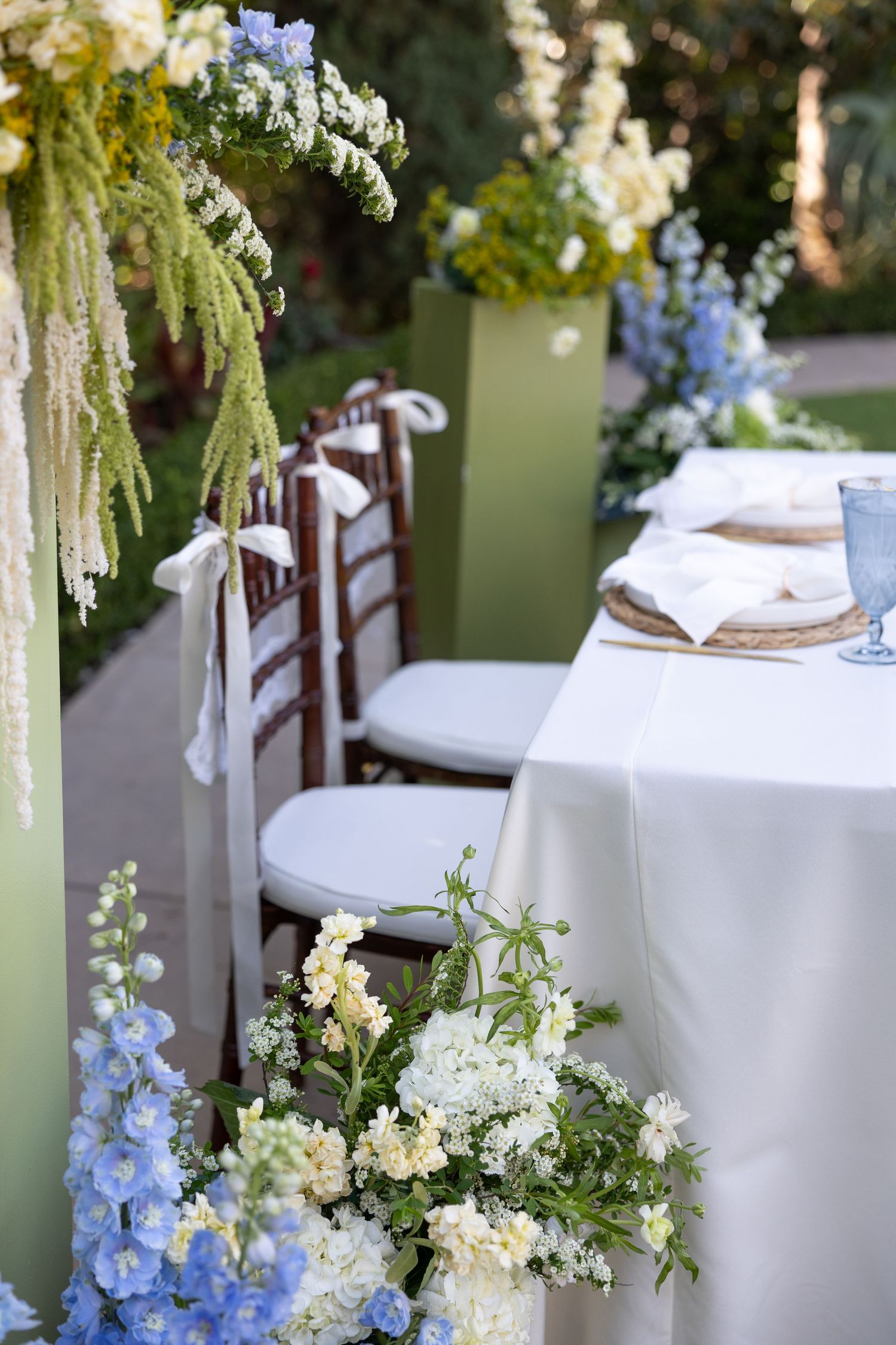 Elegant outdoor wedding reception table with blue and white floral arrangements, white tablecloth, and wooden Chiavari chairs.
