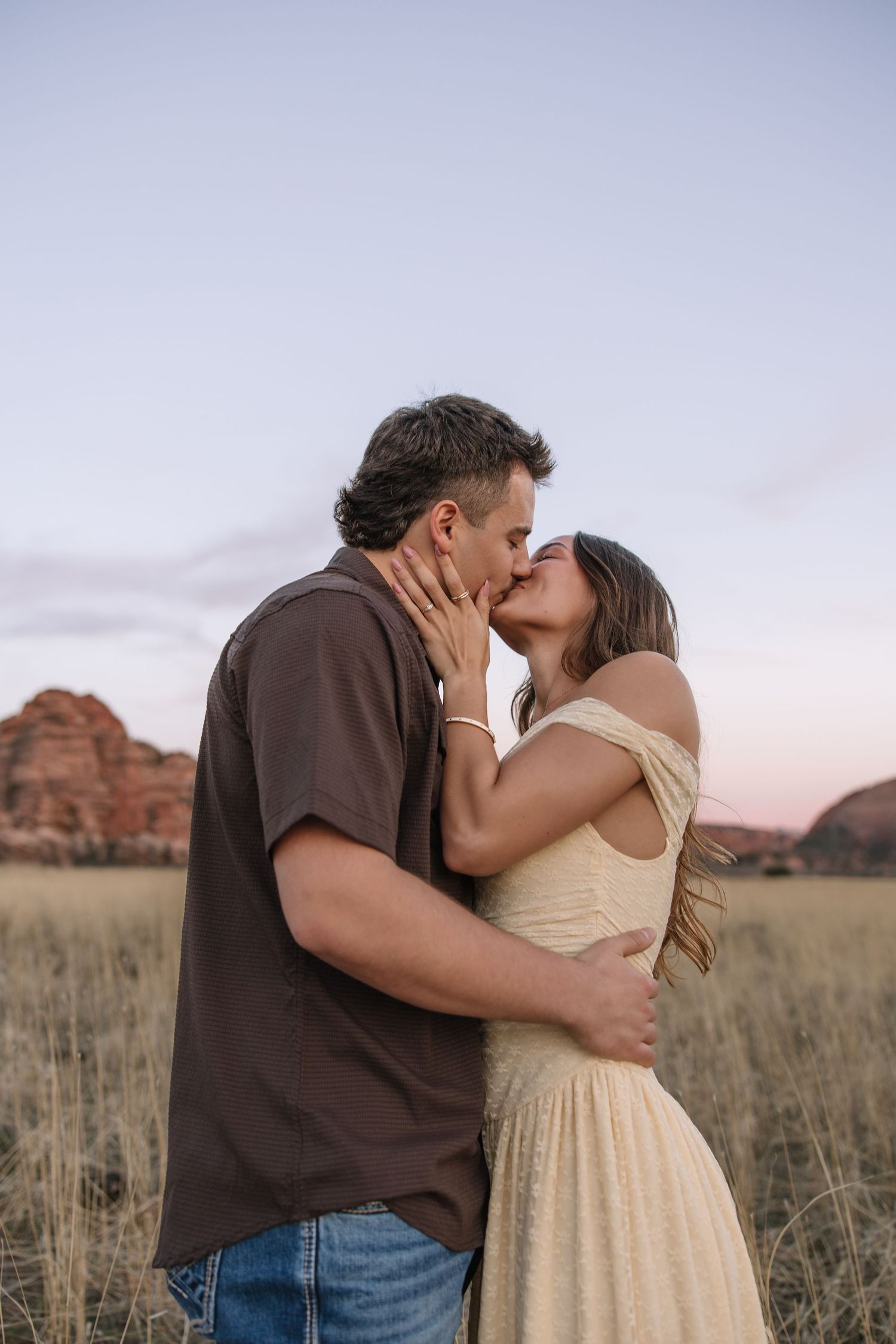 Couple sharing a romantic kiss in a golden field with red rock formations and pastel sunset sky in the background.