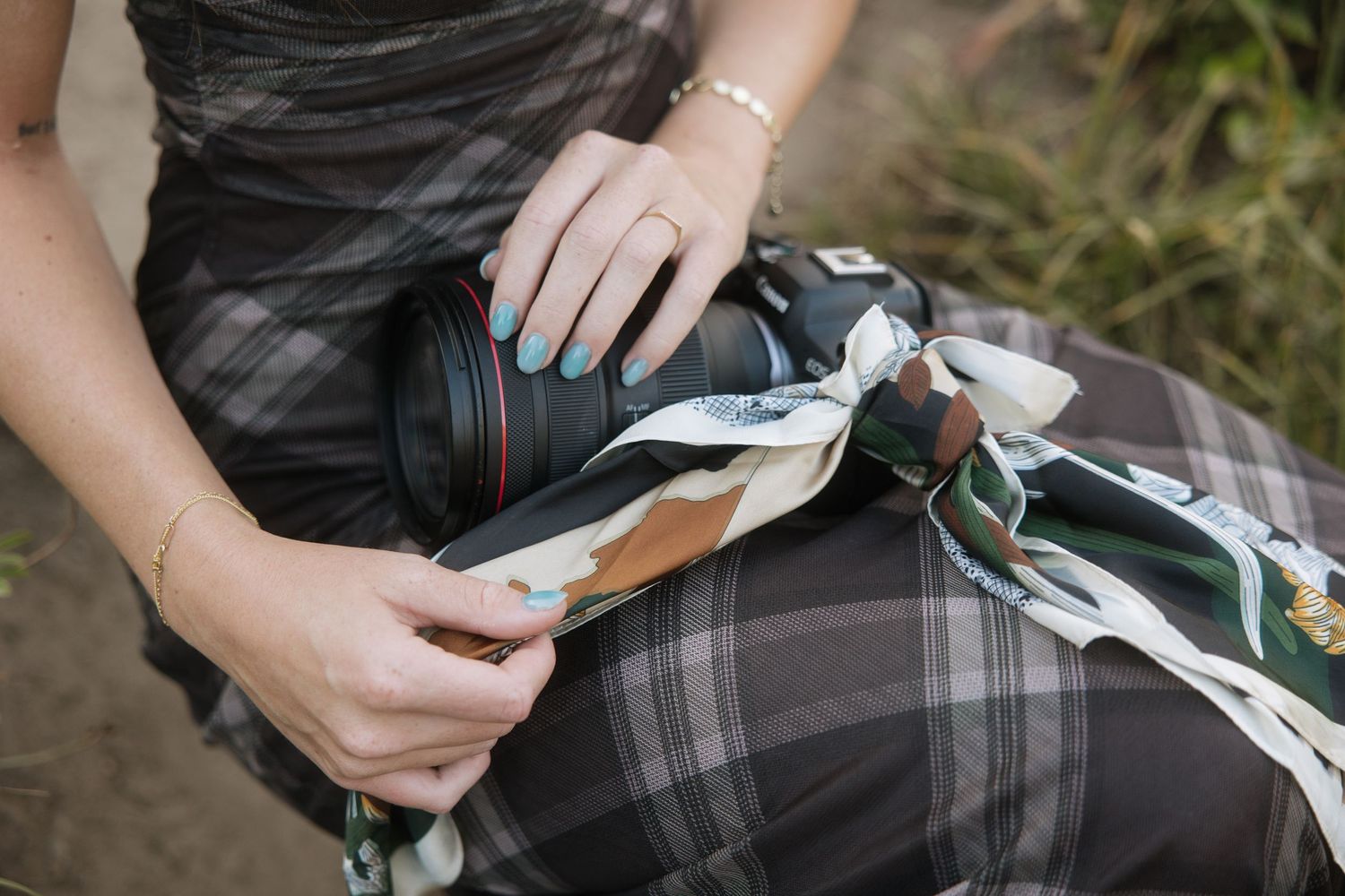 Woman in plaid dress holding a DSLR camera wrapped in a decorative floral scarf outdoors.
