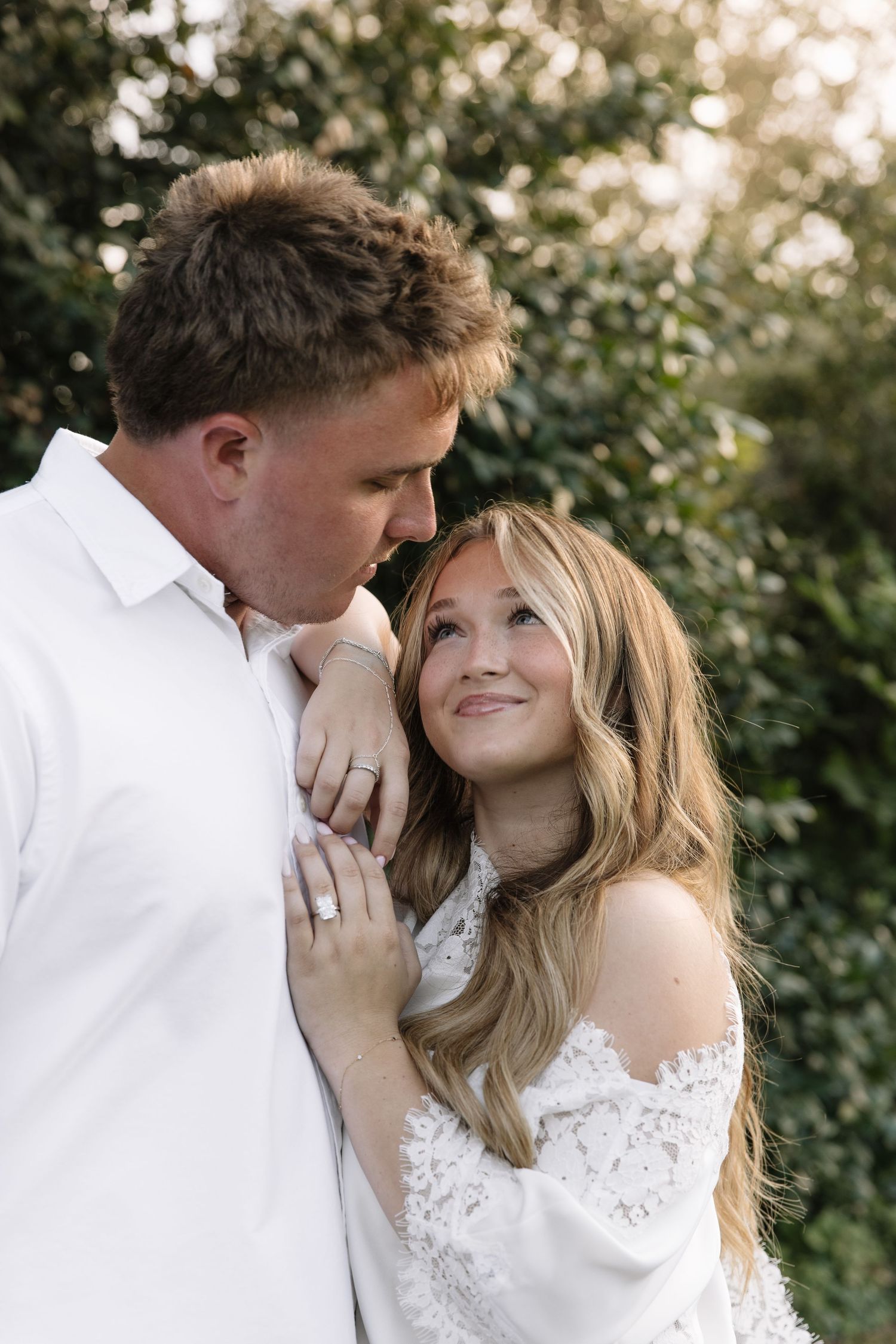 Couple in white outfits sharing a romantic moment outdoors, woman smiling up at man against lush green foliage.