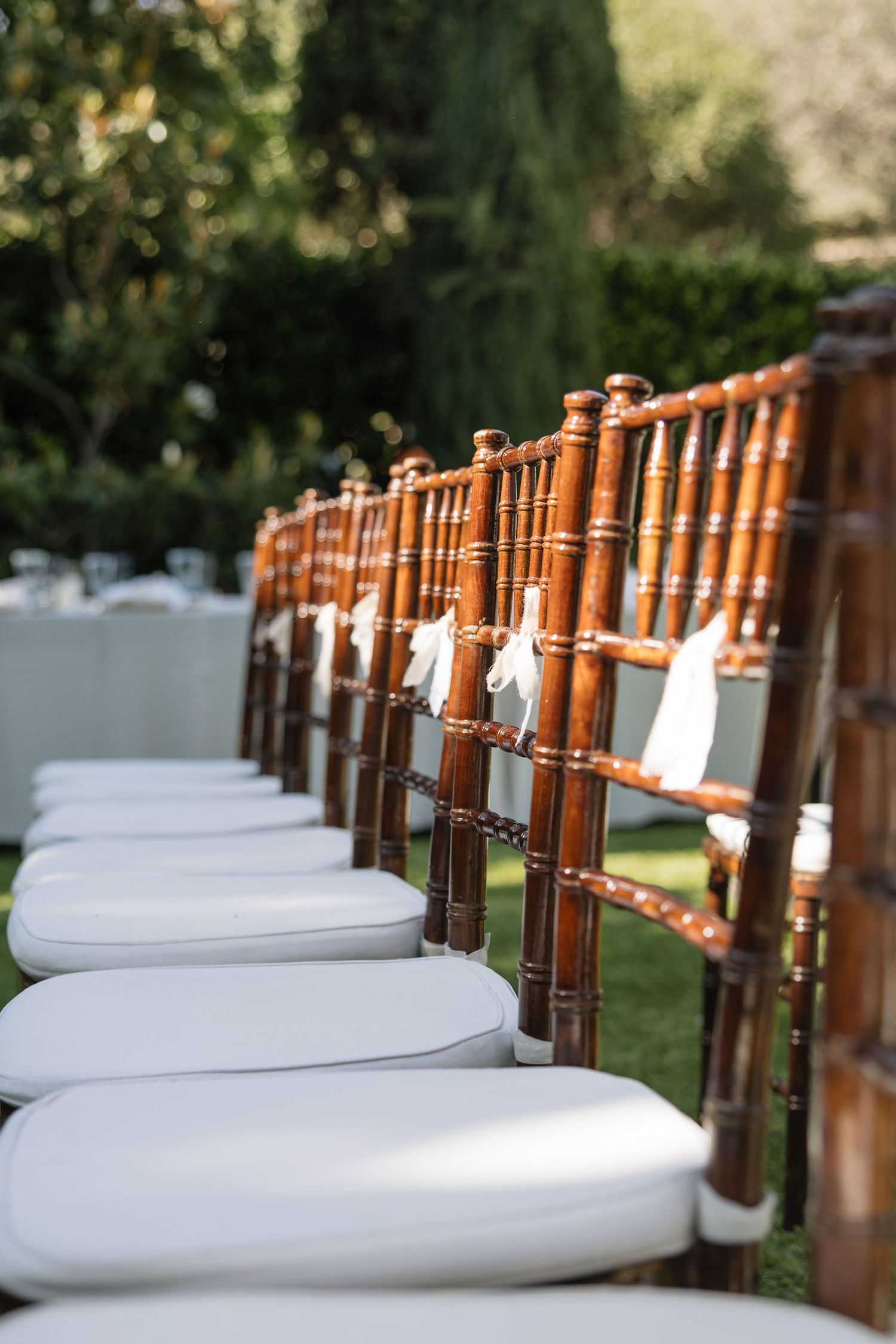 Elegant row of wooden Chiavari chairs with white cushions and ribbon bows set up outdoors for a wedding ceremony.