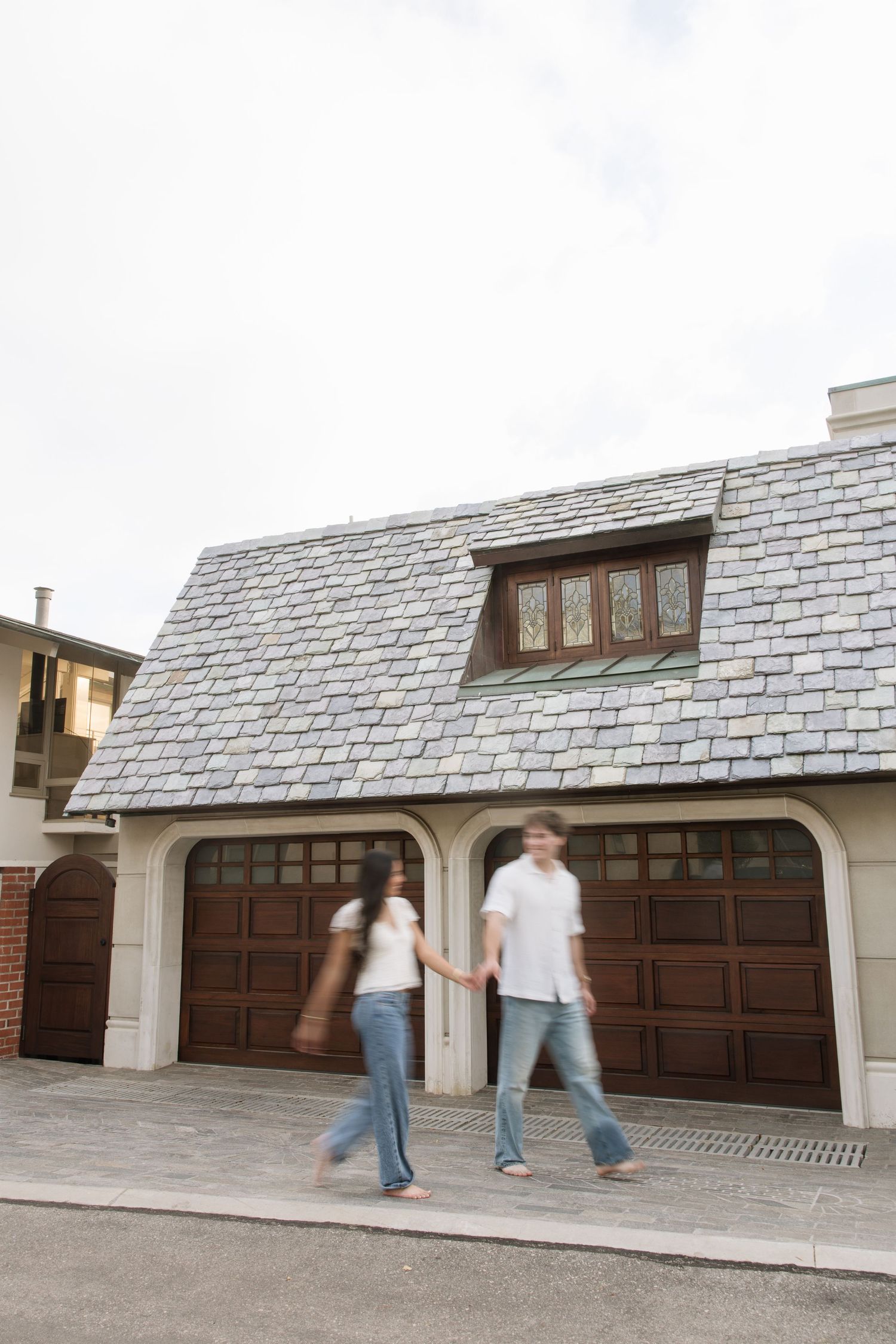 Couple holding hands walking past a charming stone-roofed garage with wooden doors and dormer window.