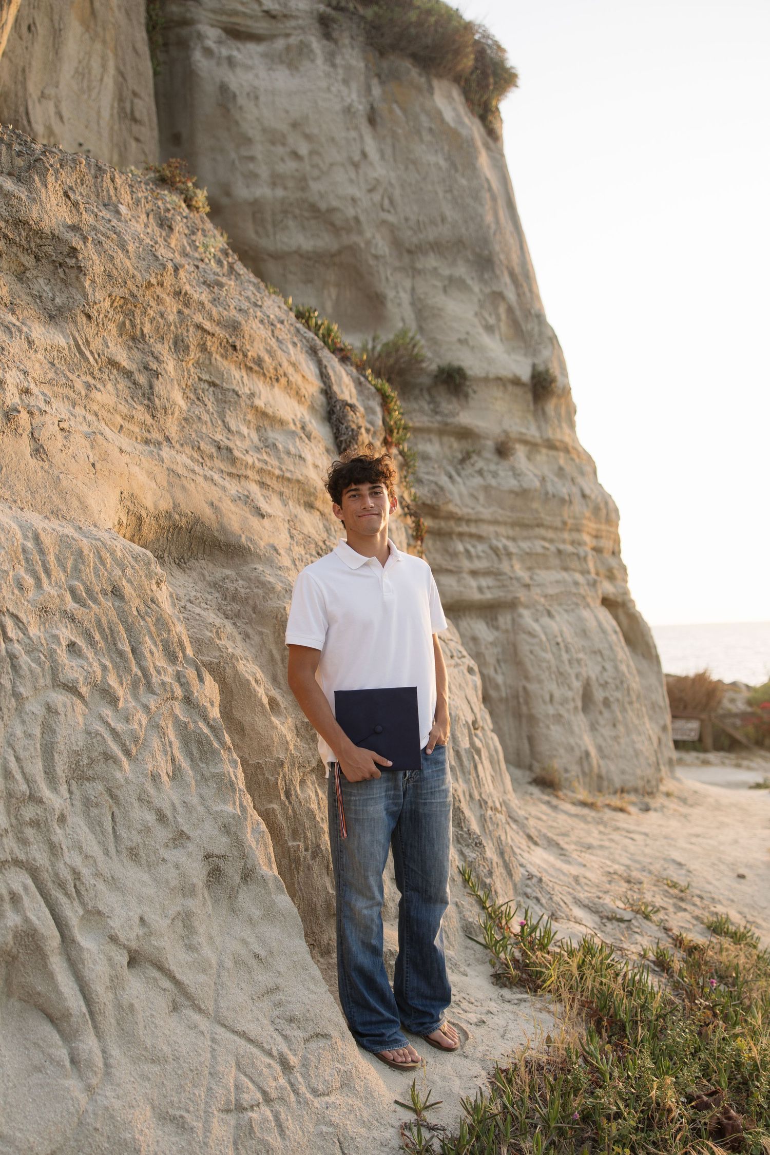 Young man in white polo and jeans standing against sandy cliffs at beach during golden hour.