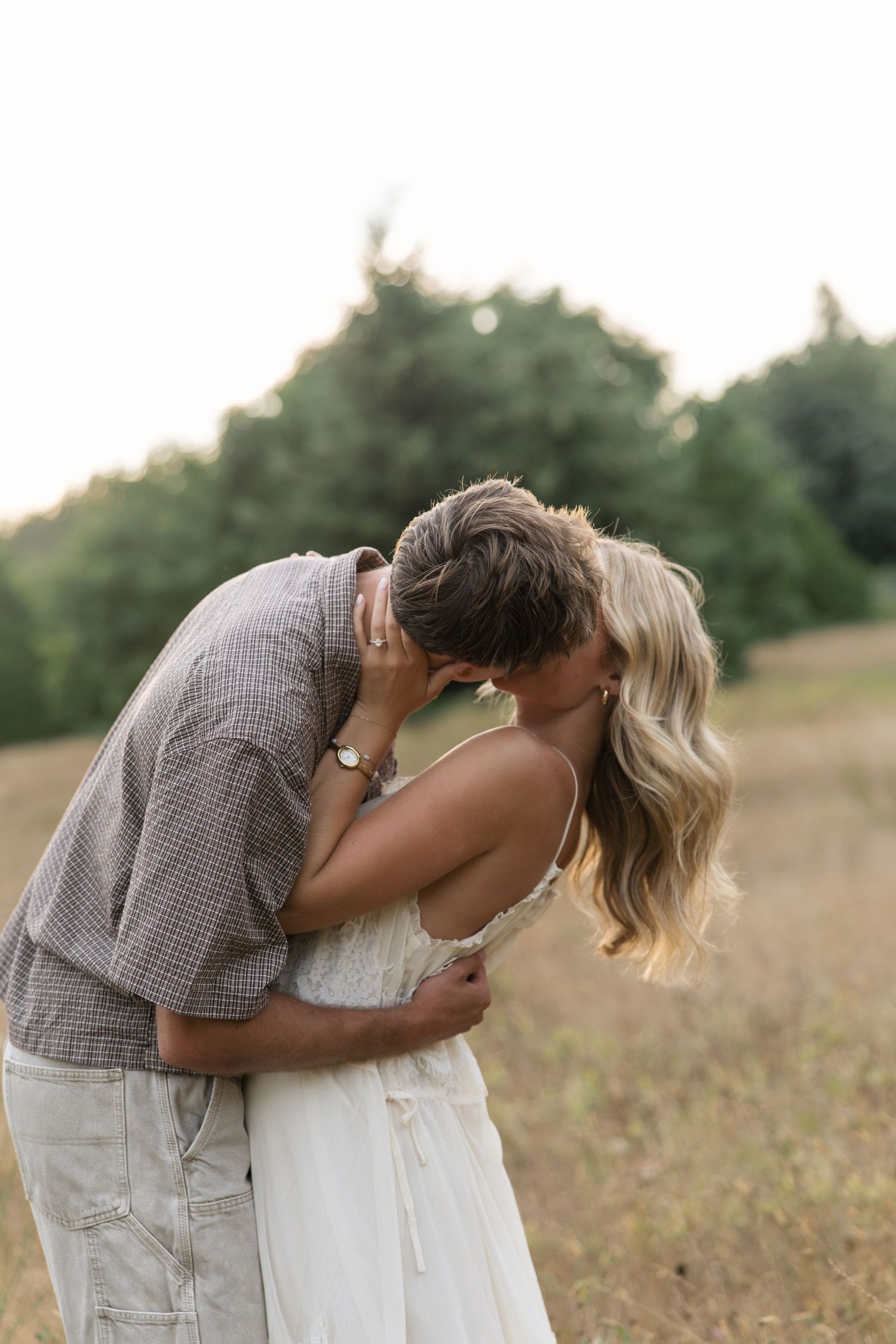 A couple sharing a romantic kiss in a golden field surrounded by lush green trees during a warm summer evening.