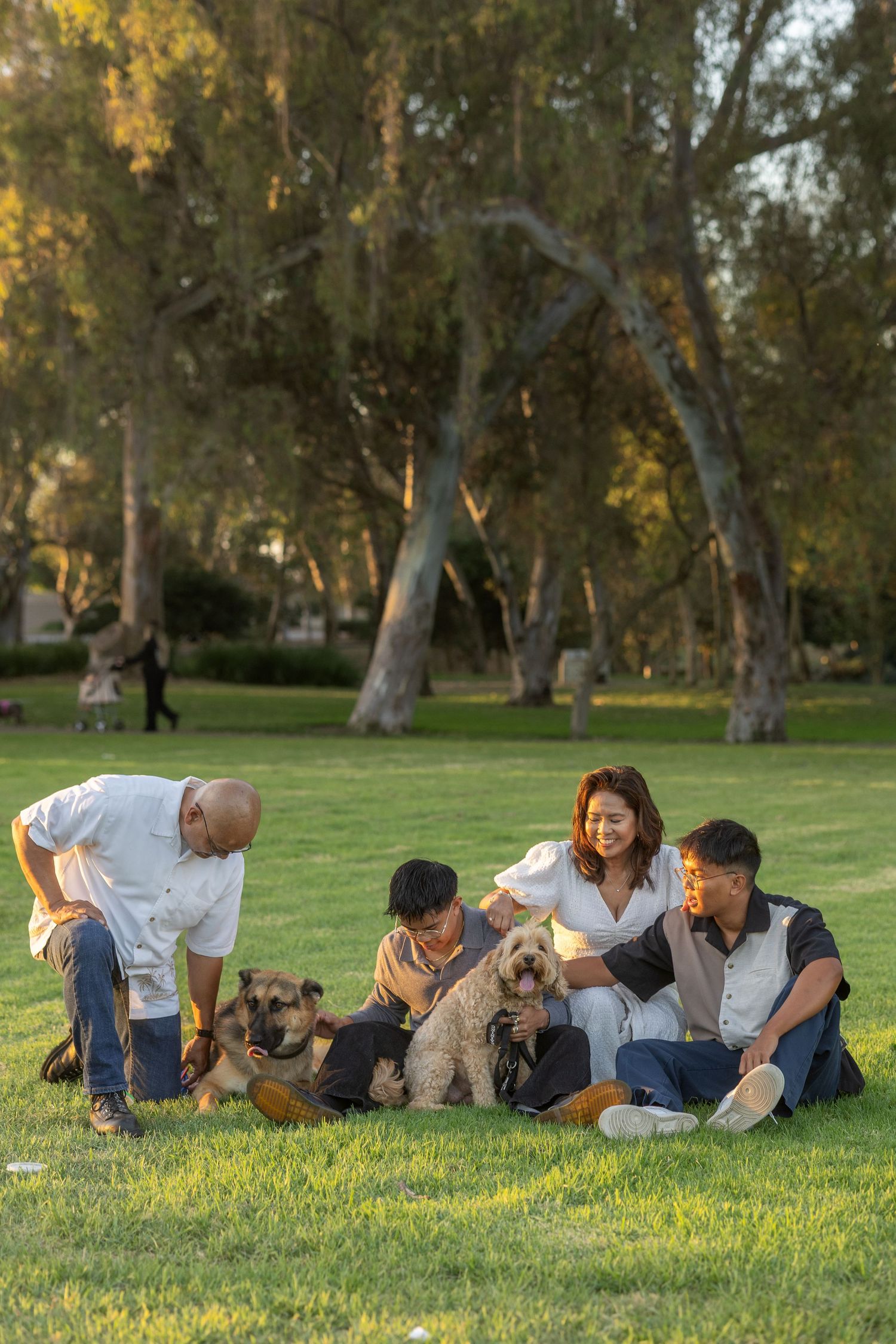 Family and dog relaxing together on grass in a park with tall trees.