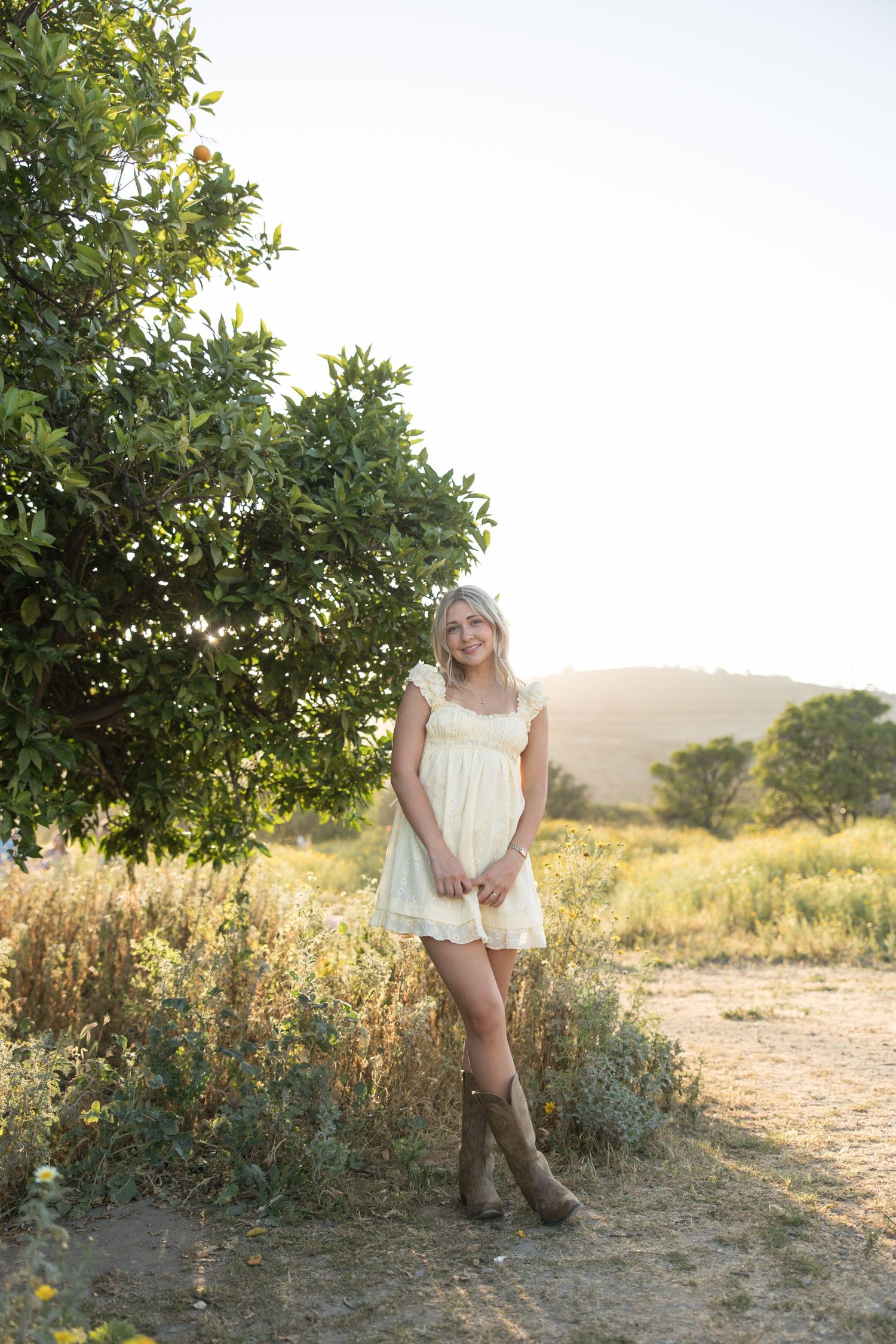 Young blonde woman in white sundress and cowboy boots standing near a tree in a golden sunlit field.
