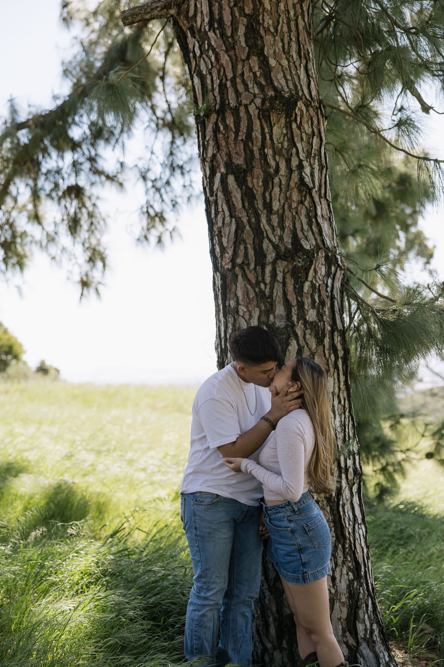 A romantic couple kissing against a pine tree in a lush green meadow, both wearing casual jeans and white tops.