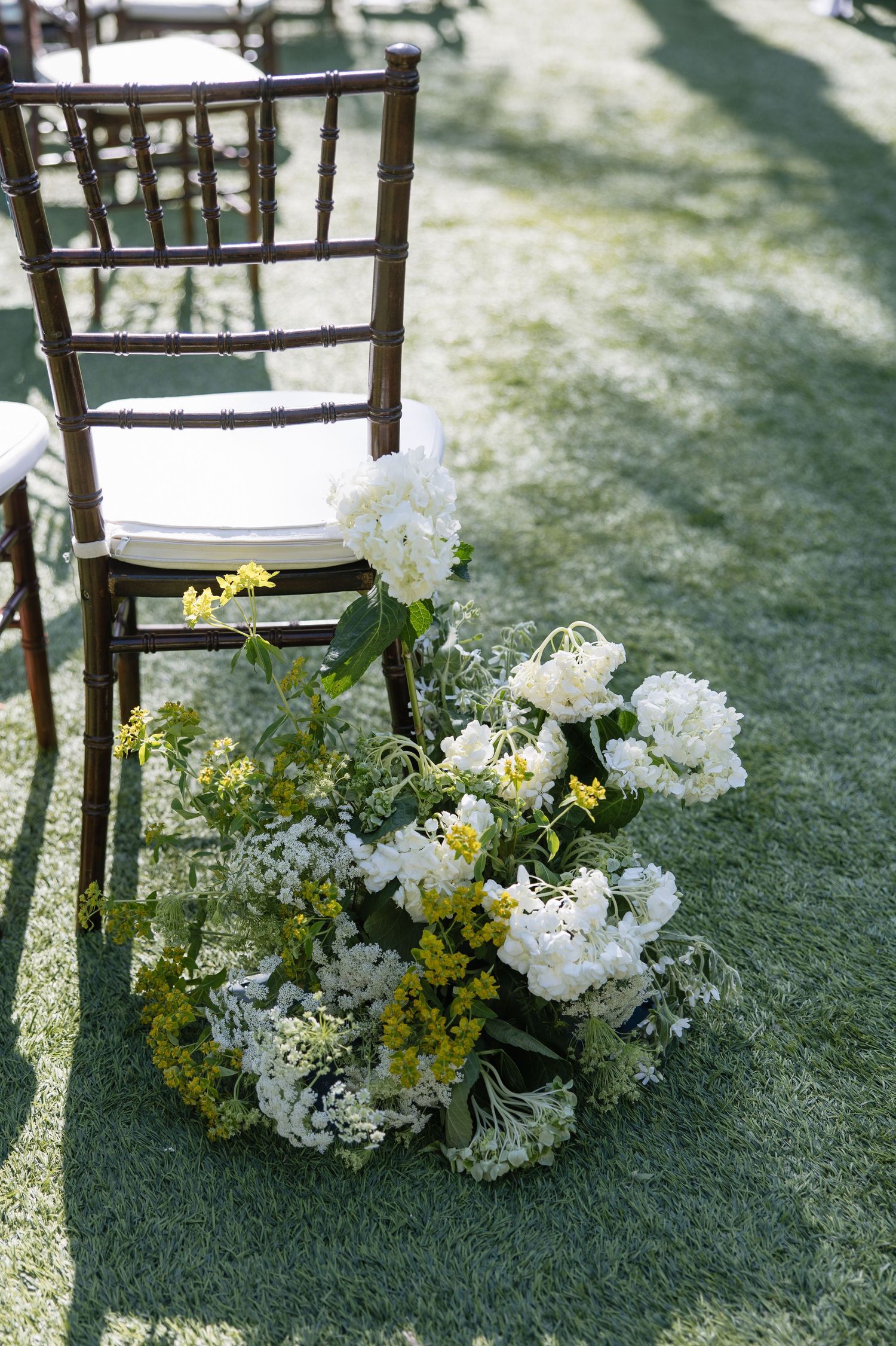 Outdoor wedding ceremony aisle with white peonies and yellow floral arrangement beside dark wood Chiavari chair on green grass.