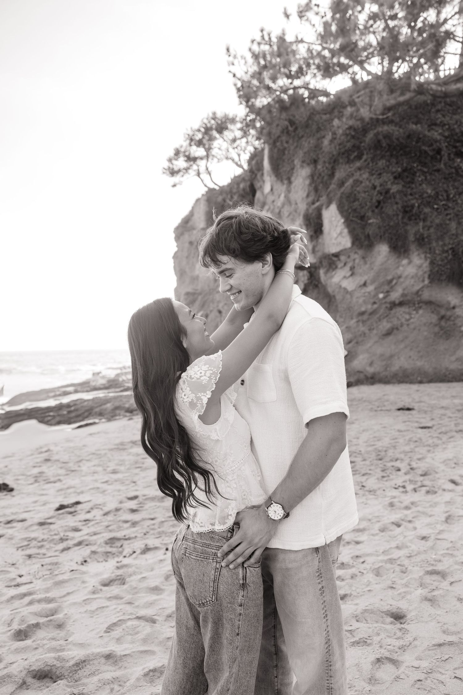 Black and white photo of a romantic couple embracing on a sandy beach with rocky cliffs in the background.
