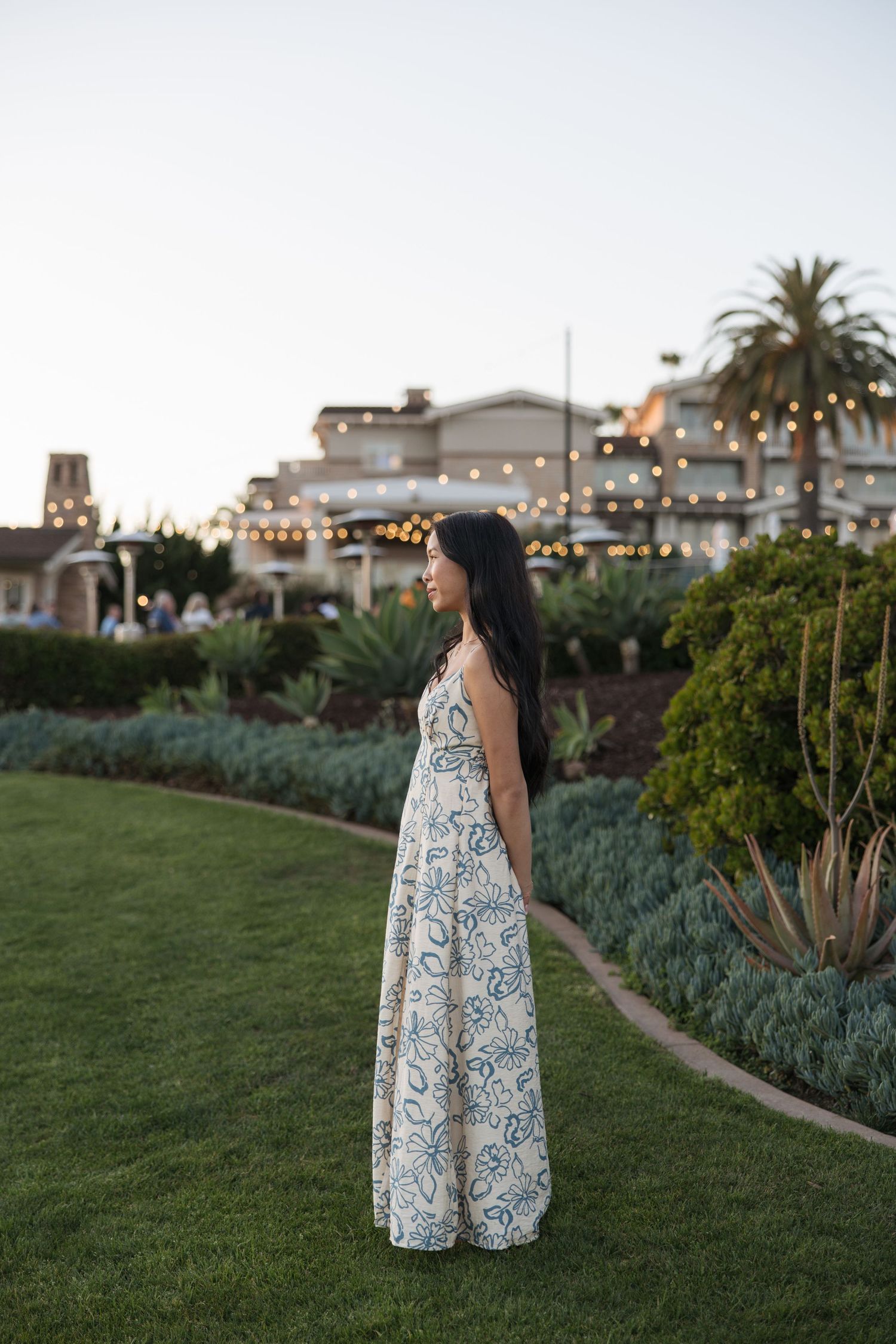 Woman in floral maxi dress standing on green lawn at dusk with string lights and palm trees in background.