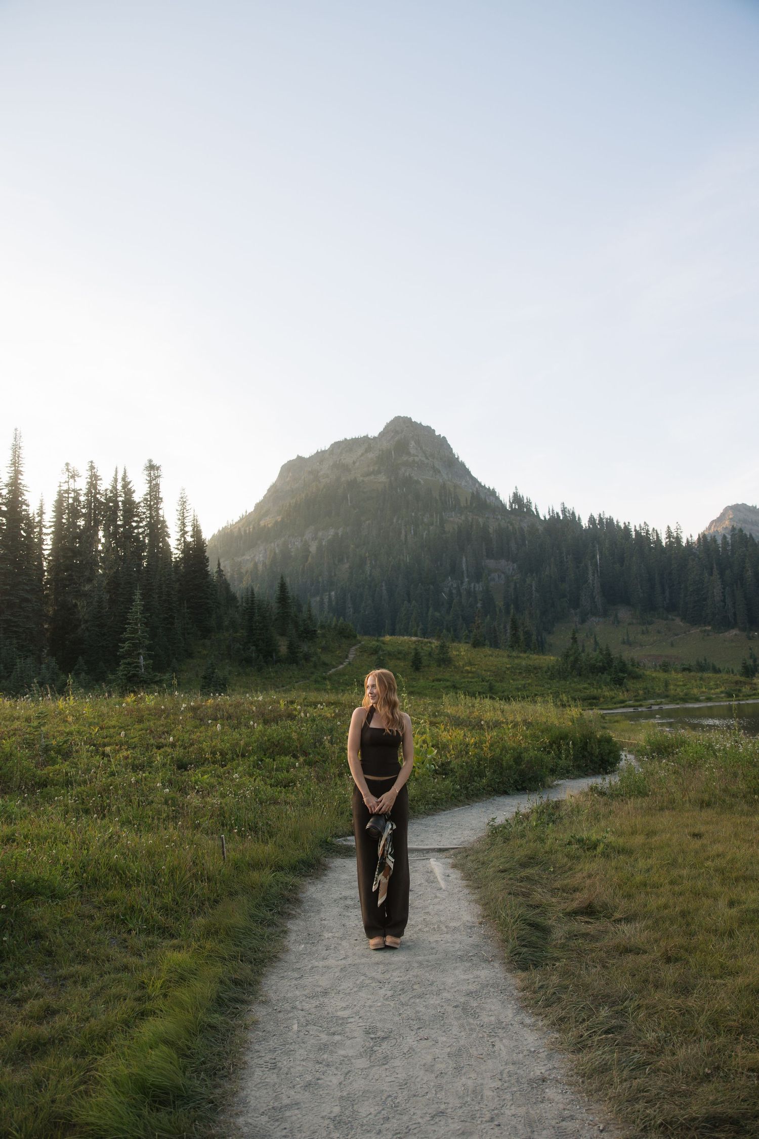 Woman in black outfit standing on a winding path with evergreen trees and mountain backdrop at golden hour.
