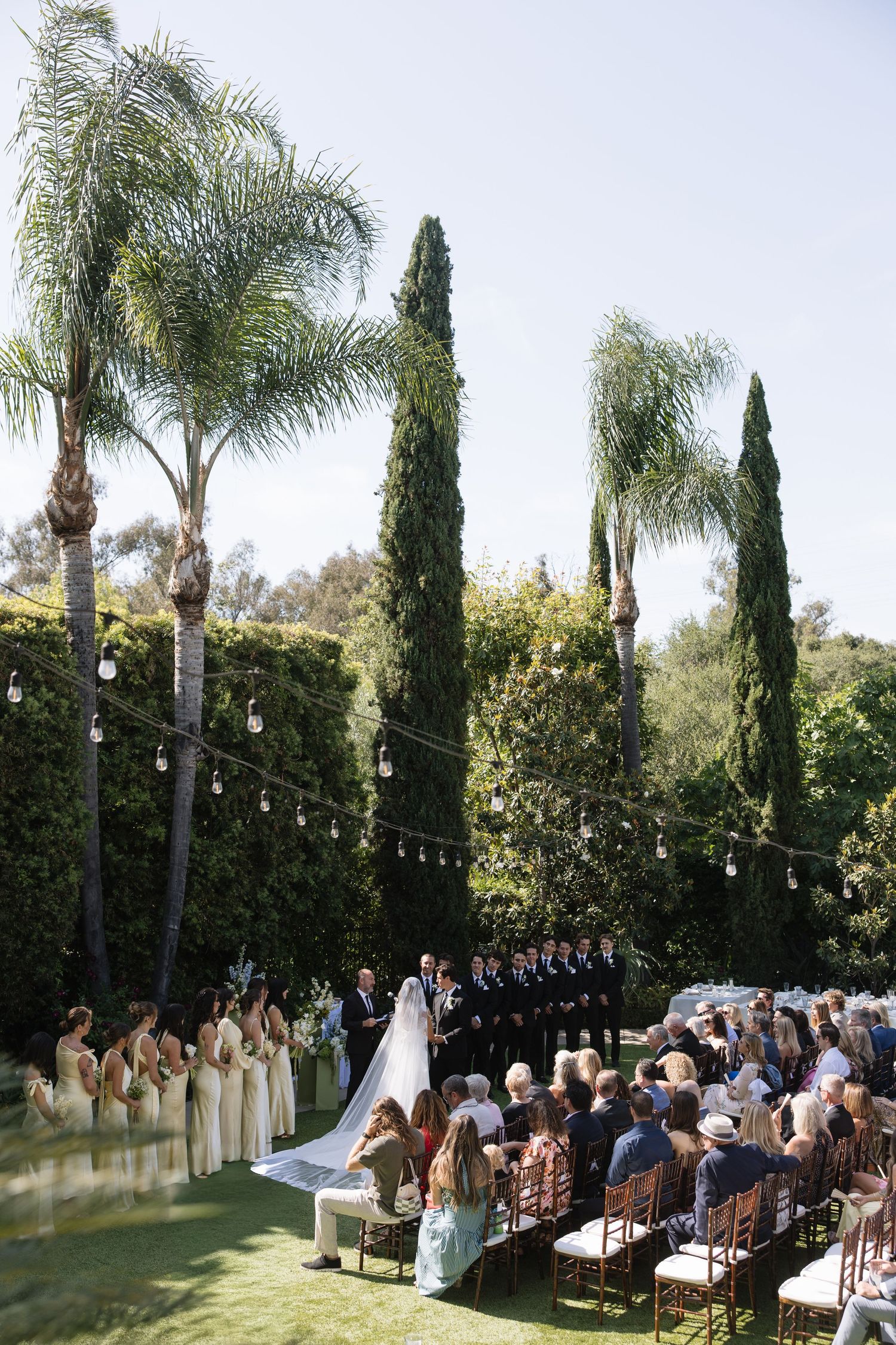 Outdoor wedding ceremony on lush green lawn with palm trees, string lights, bridesmaids, and seated guests in warm sunlight.