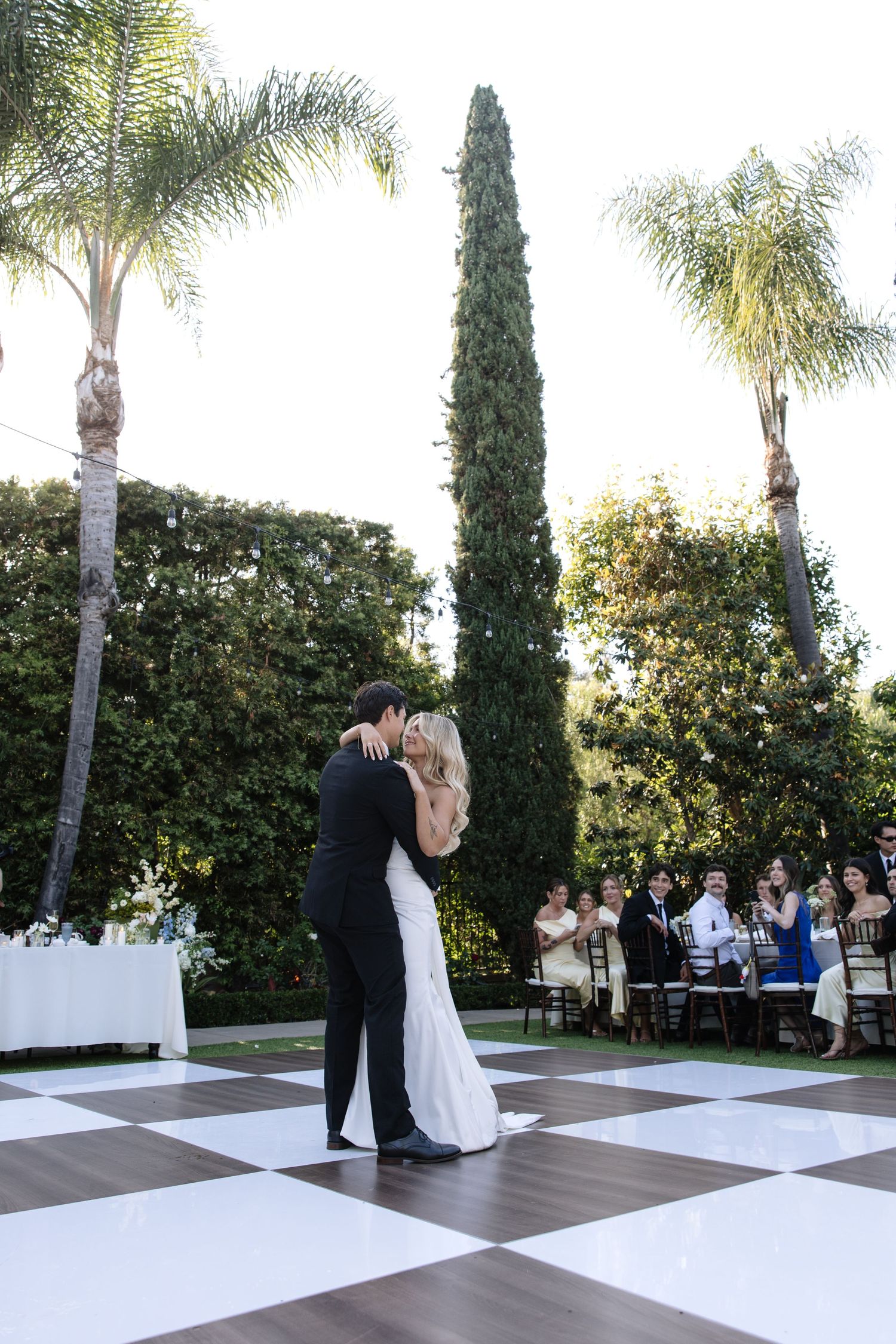 Bride and groom share first dance on checkered floor outdoors surrounded by palm trees and wedding guests.