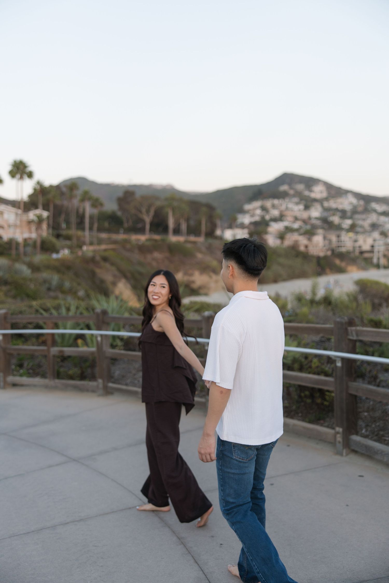 Smiling couple walking hand in hand along a seaside path with a wooden railing, palm trees, and hillside homes at sunset.