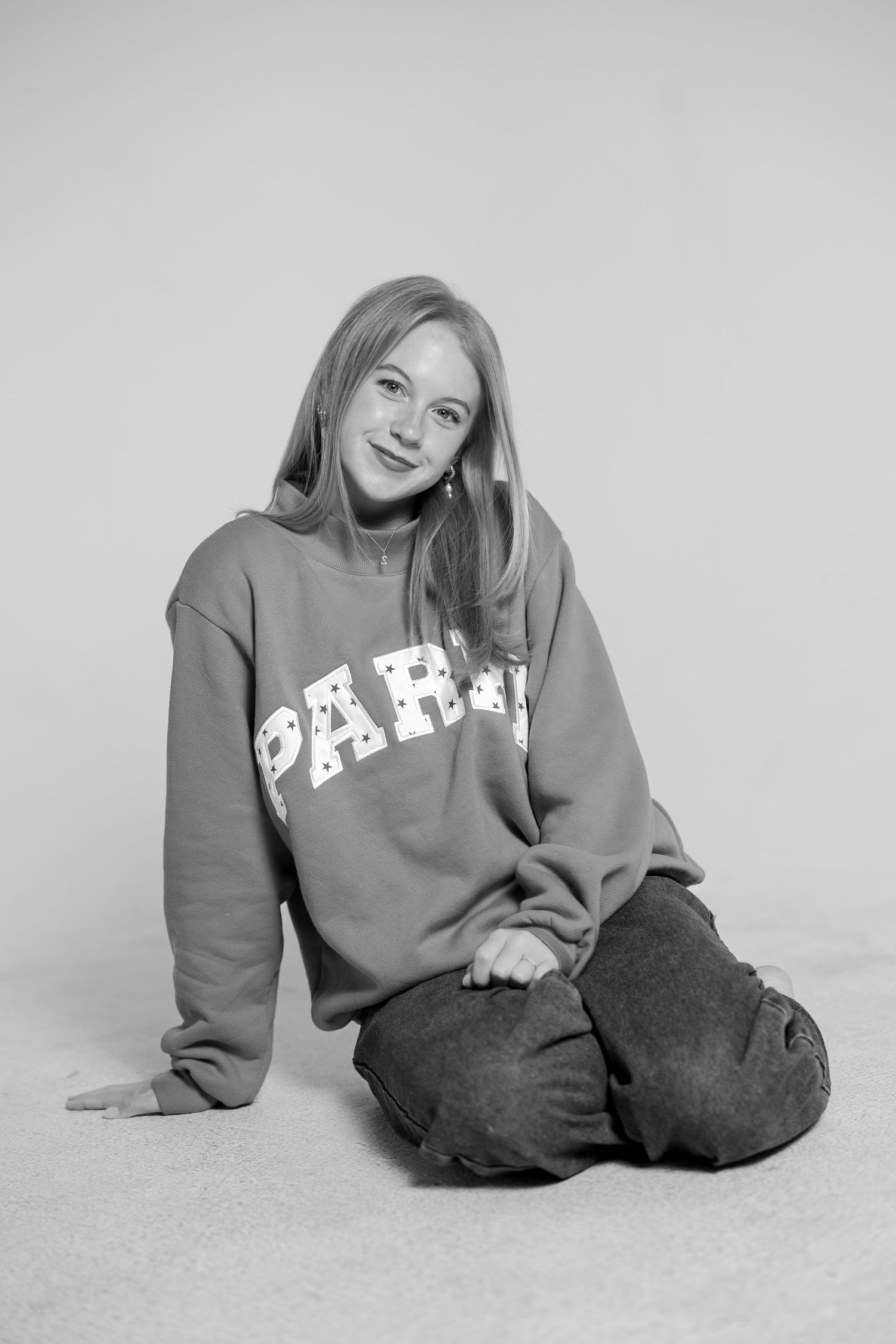Young woman sitting on floor wearing a Paris sweatshirt and jeans, smiling in a black and white studio photo.