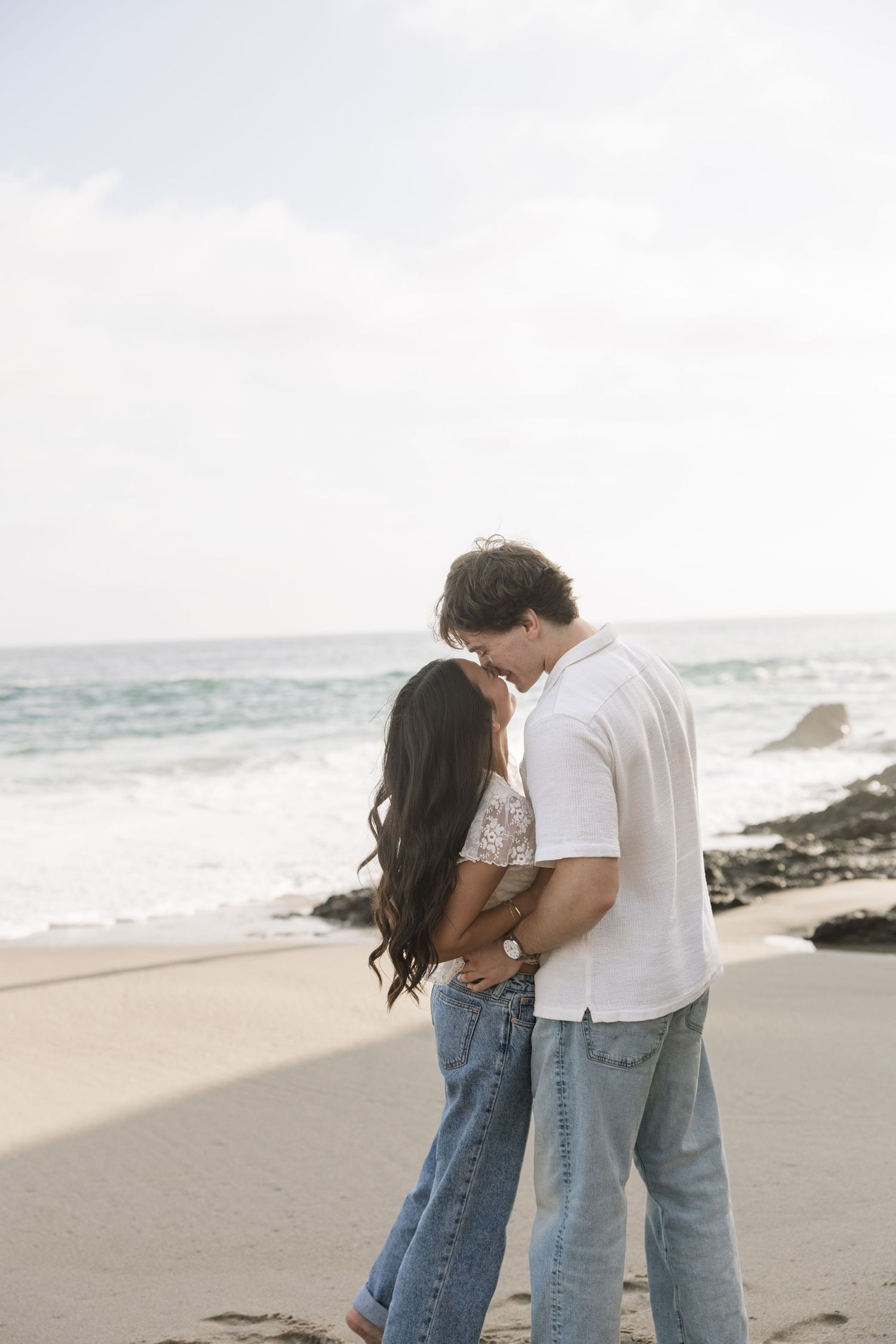 A couple shares a romantic moment kissing on a sandy beach with ocean waves in the background.