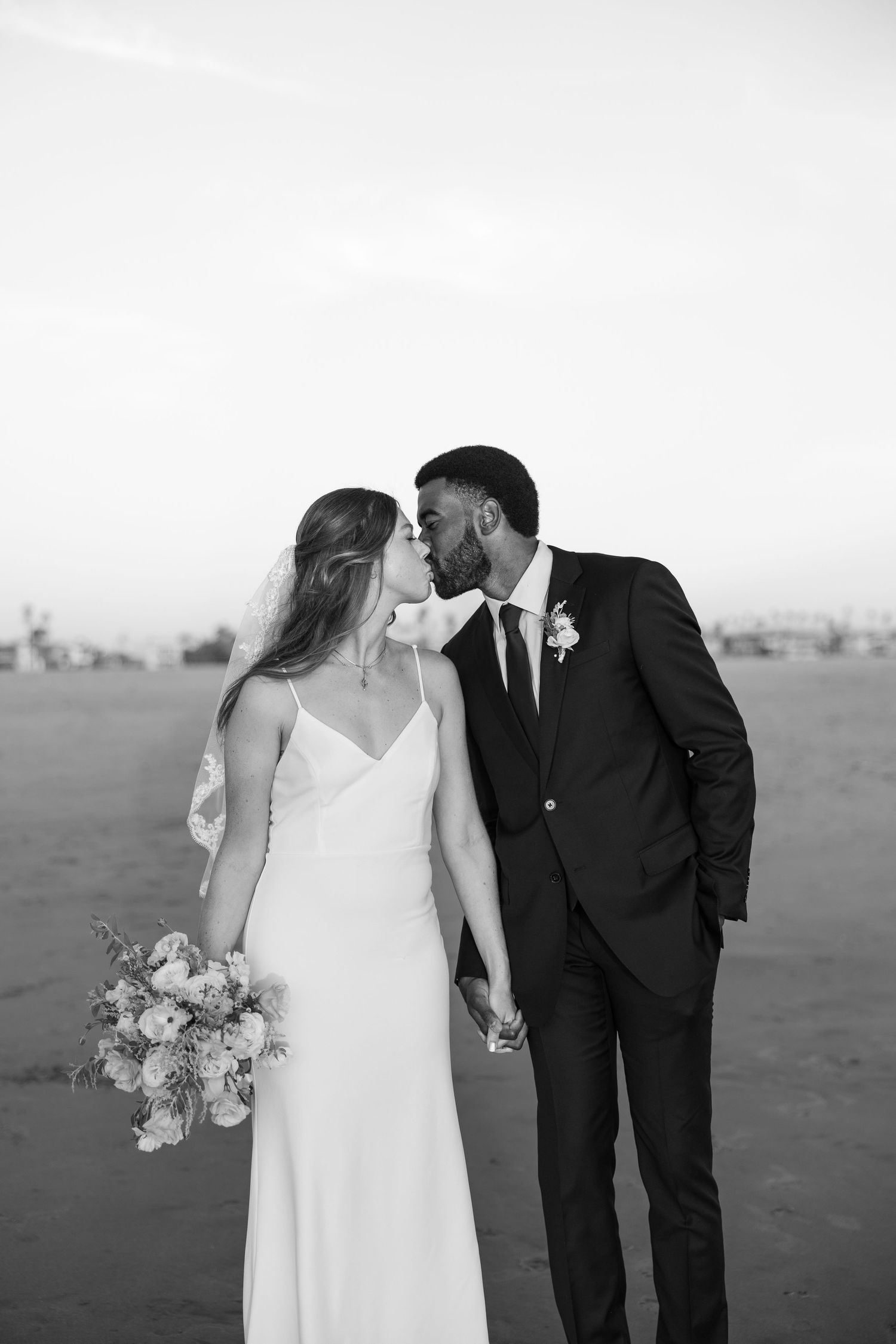 A romantic wedding couple shares a kiss on the beach in black and white photography.
