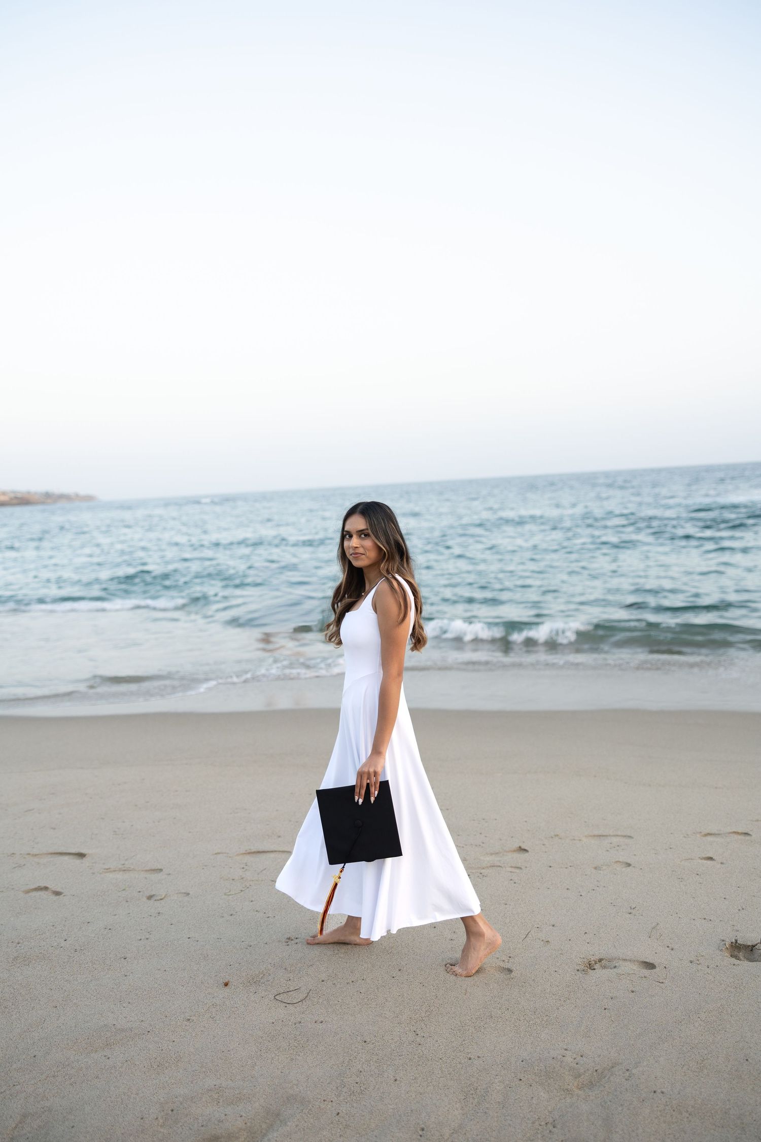 Woman in white maxi dress walking barefoot on sandy beach holding black clutch with ocean in background.