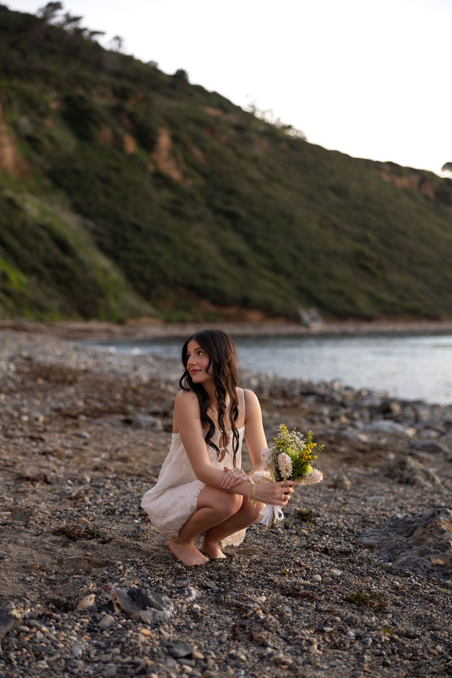 A person in flowing white dress crouches to pick flowers on a rocky beach.