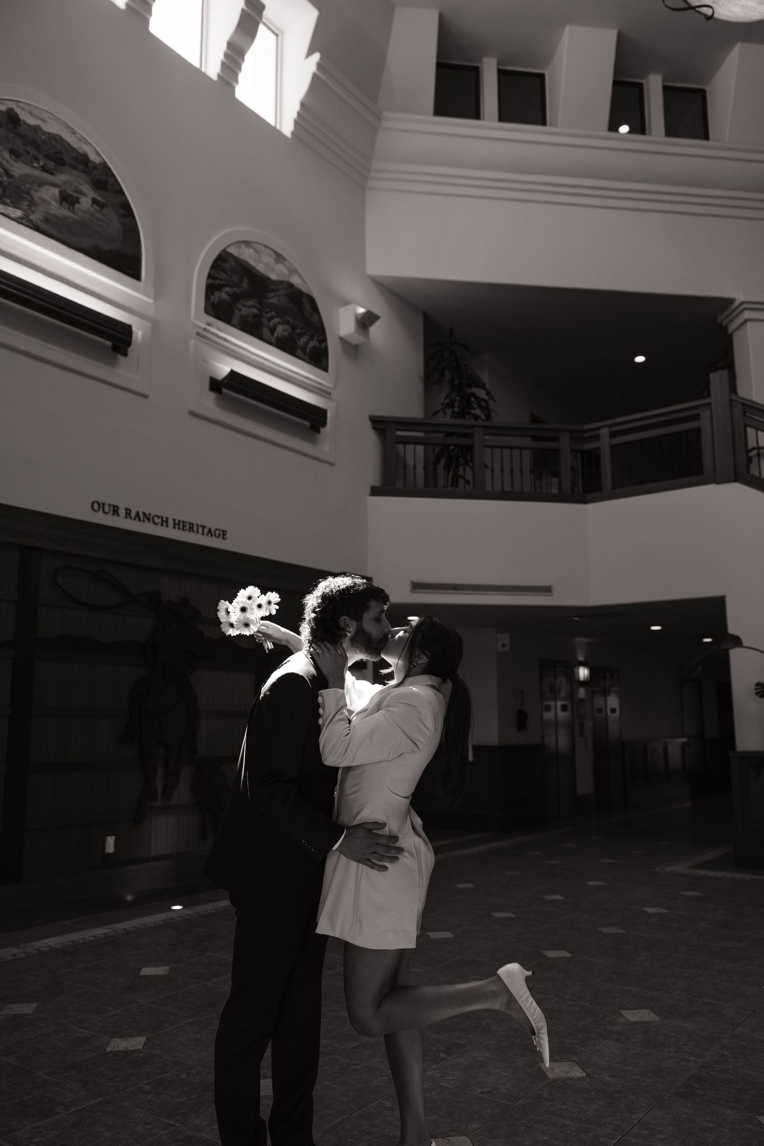 Black and white photo of a couple kissing in a grand lobby, the woman in a short white dress kicking up her heel and holding a bouquet.