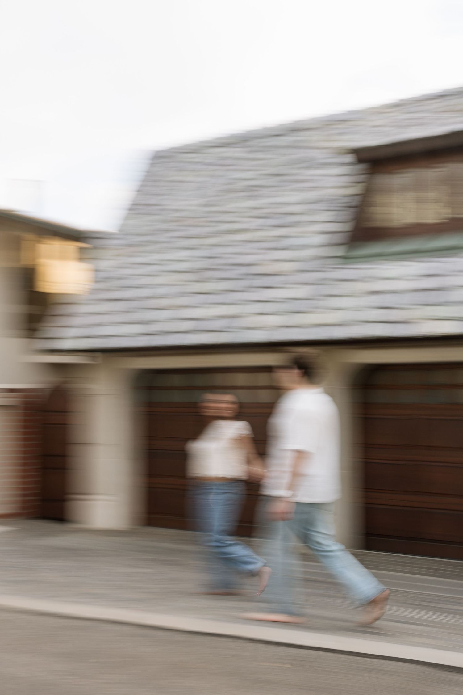 Blurred motion shot of a couple holding hands walking past a house with a slate roof and wooden garage doors.