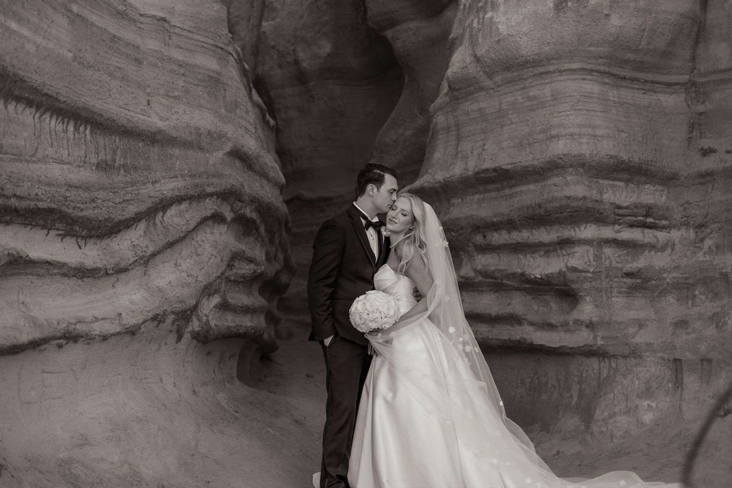 Black and white photo of a bride and groom sharing a kiss inside a stunning sandstone canyon on their wedding day.