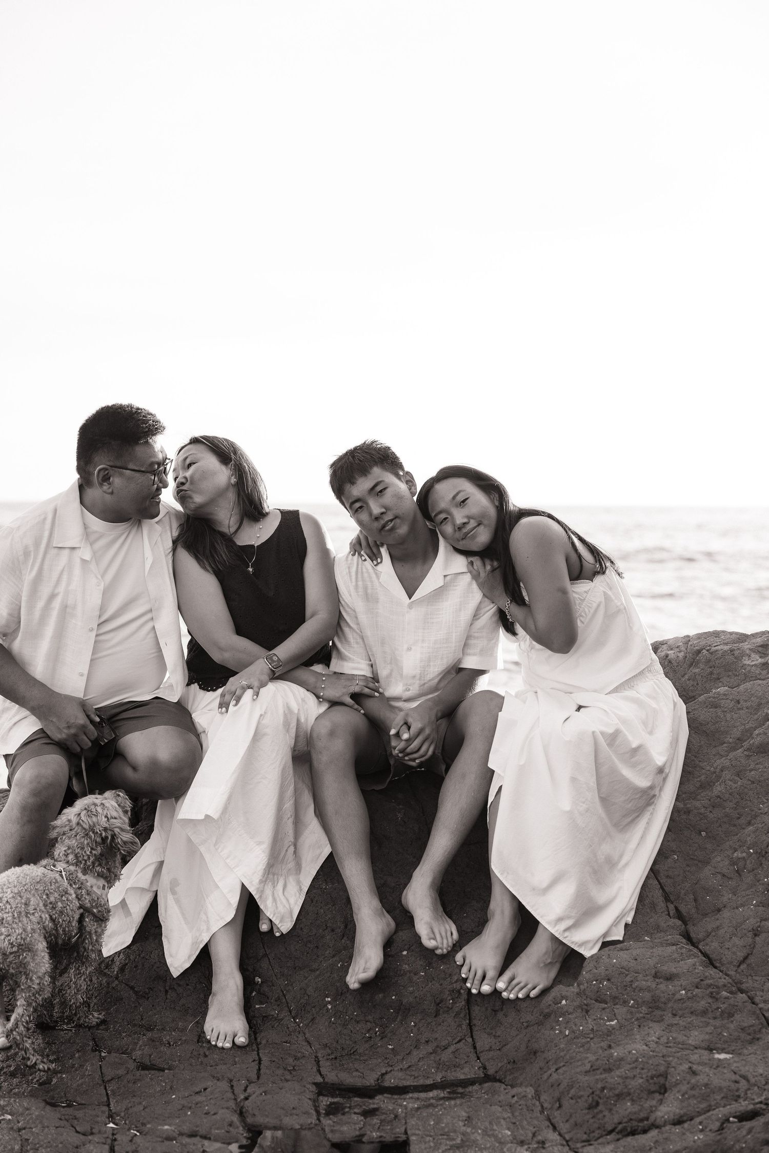 Black and white photo of four people sitting on coastal rocks near the ocean, leaning on each other with a small dog nearby.
