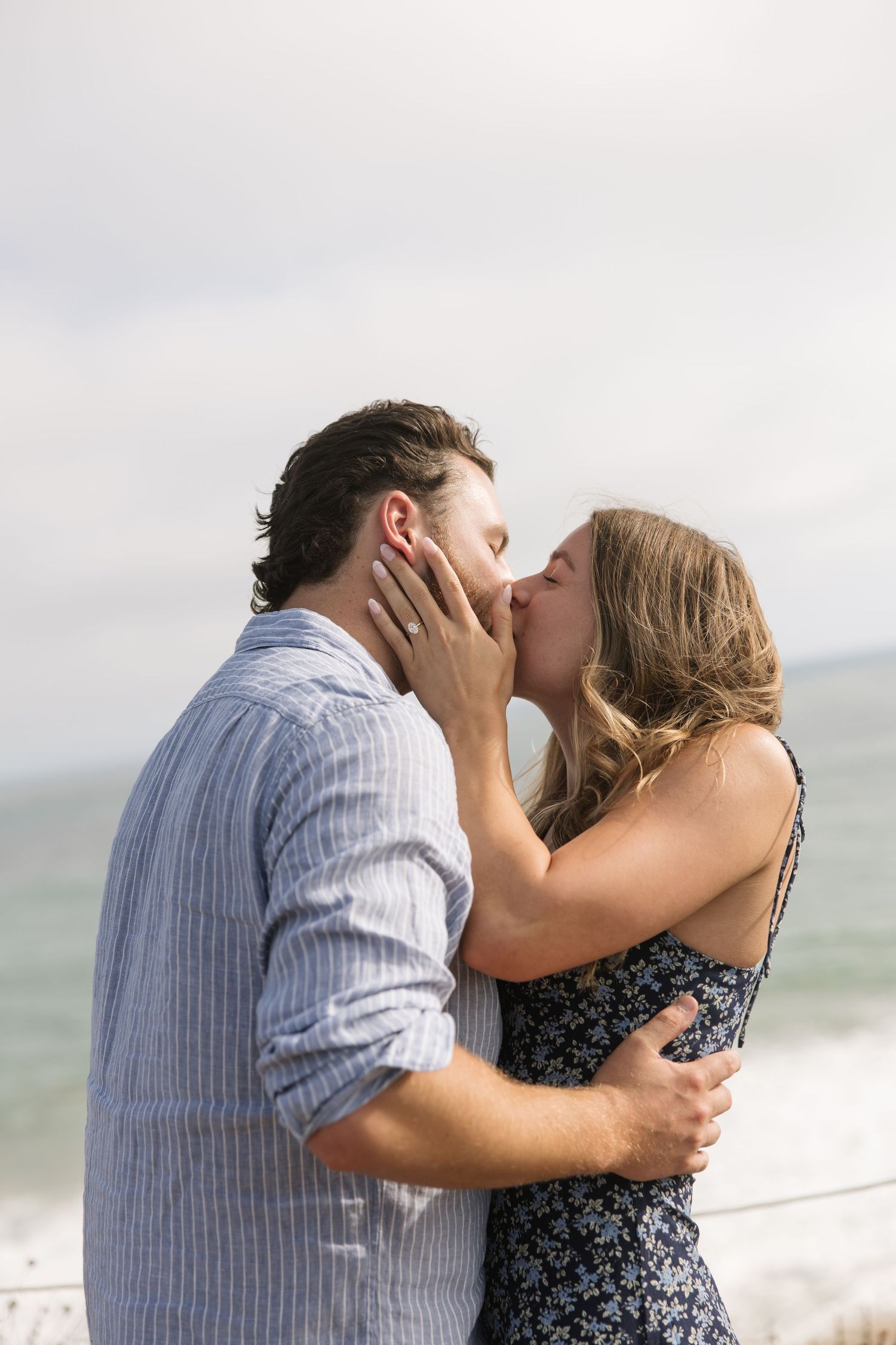 Couple sharing a romantic kiss on the beach, man in blue striped shirt embracing woman in floral dress by the ocean.