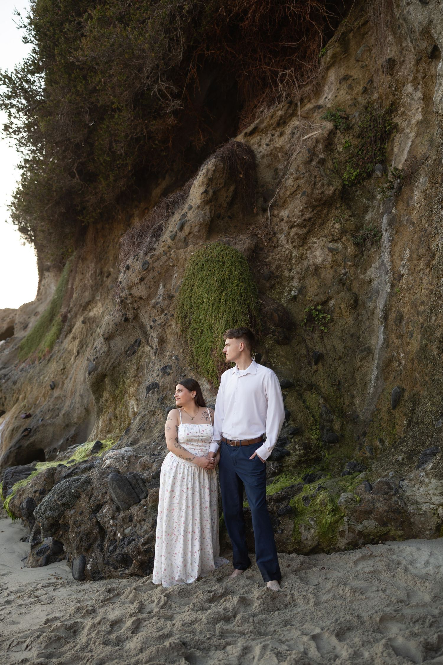 Couple standing together on sandy beach against rocky cliff, woman in white dress and man in white shirt and navy pants.