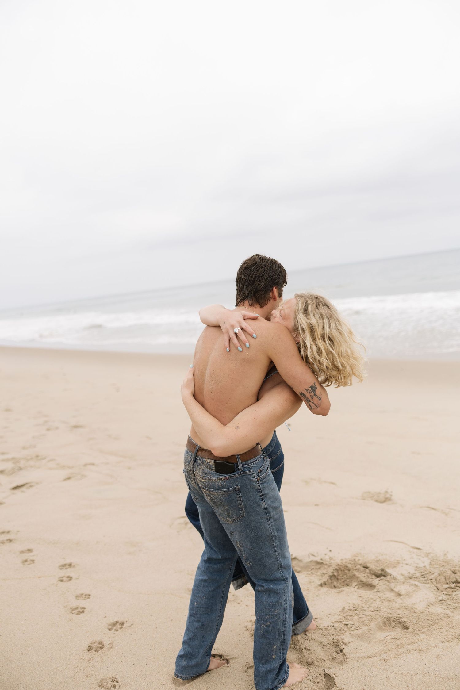 Romantic couple embracing and kissing on a sandy beach, man shirtless in jeans, woman in jeans, ocean waves in background.