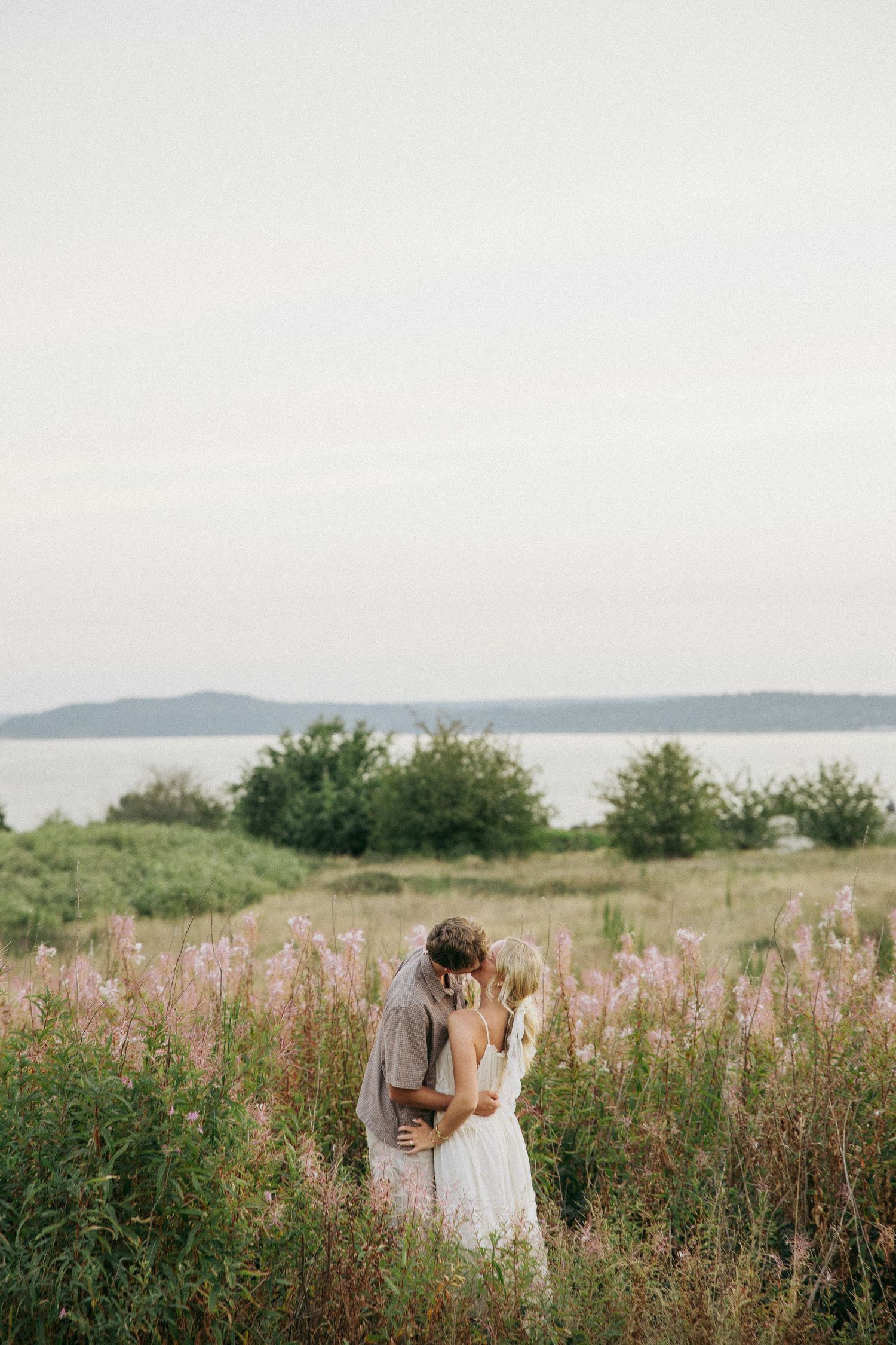 A bride and groom share a romantic kiss in a wildflower field with a serene lake and hills in the background.