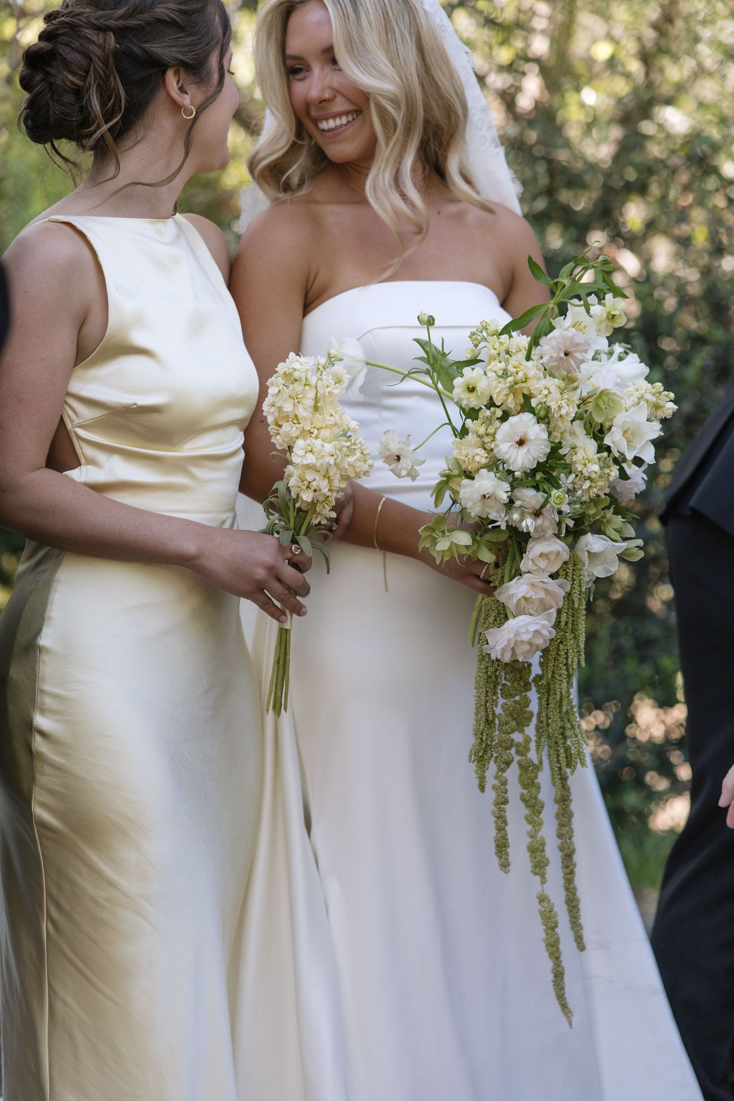 Smiling bride in white strapless gown holds cascading bouquet beside bridesmaid in champagne satin dress outdoors.