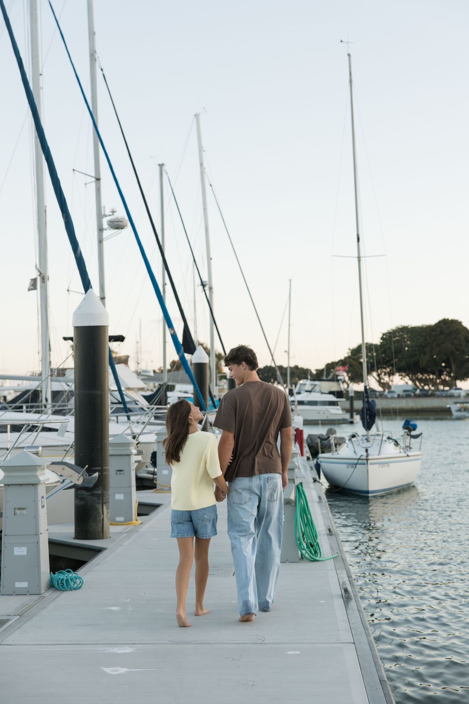 Barefoot couple walking hand in hand along a marina dock lined with sailboats and tall masts at sunset.
