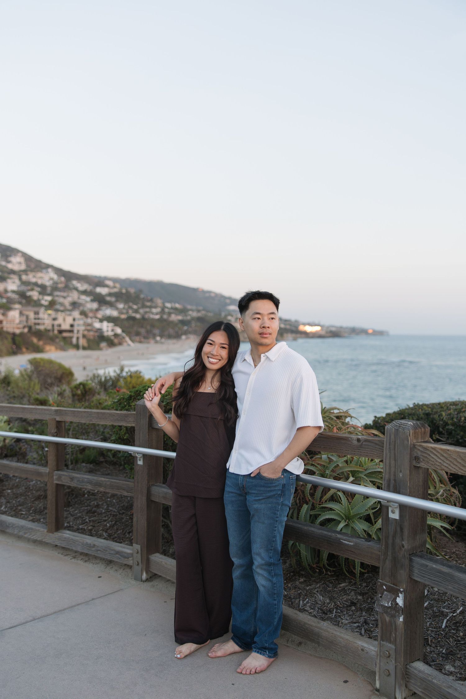 Smiling couple standing barefoot at a wooden railing overlooking a scenic coastal beach and hillside homes at sunset.