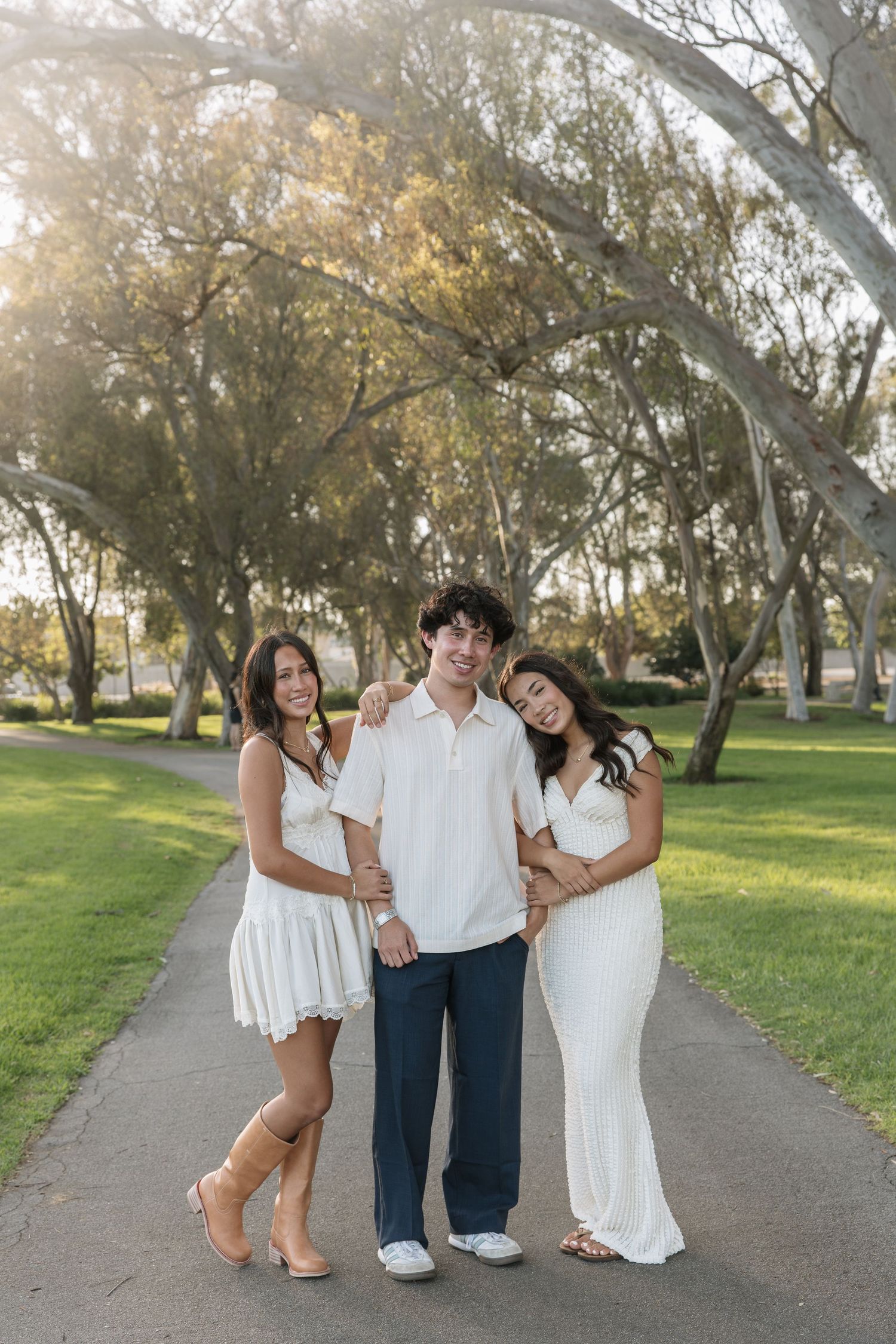 Three young people in white and navy outfits pose arm in arm on a paved park path lined with tall eucalyptus trees in warm sunlight.