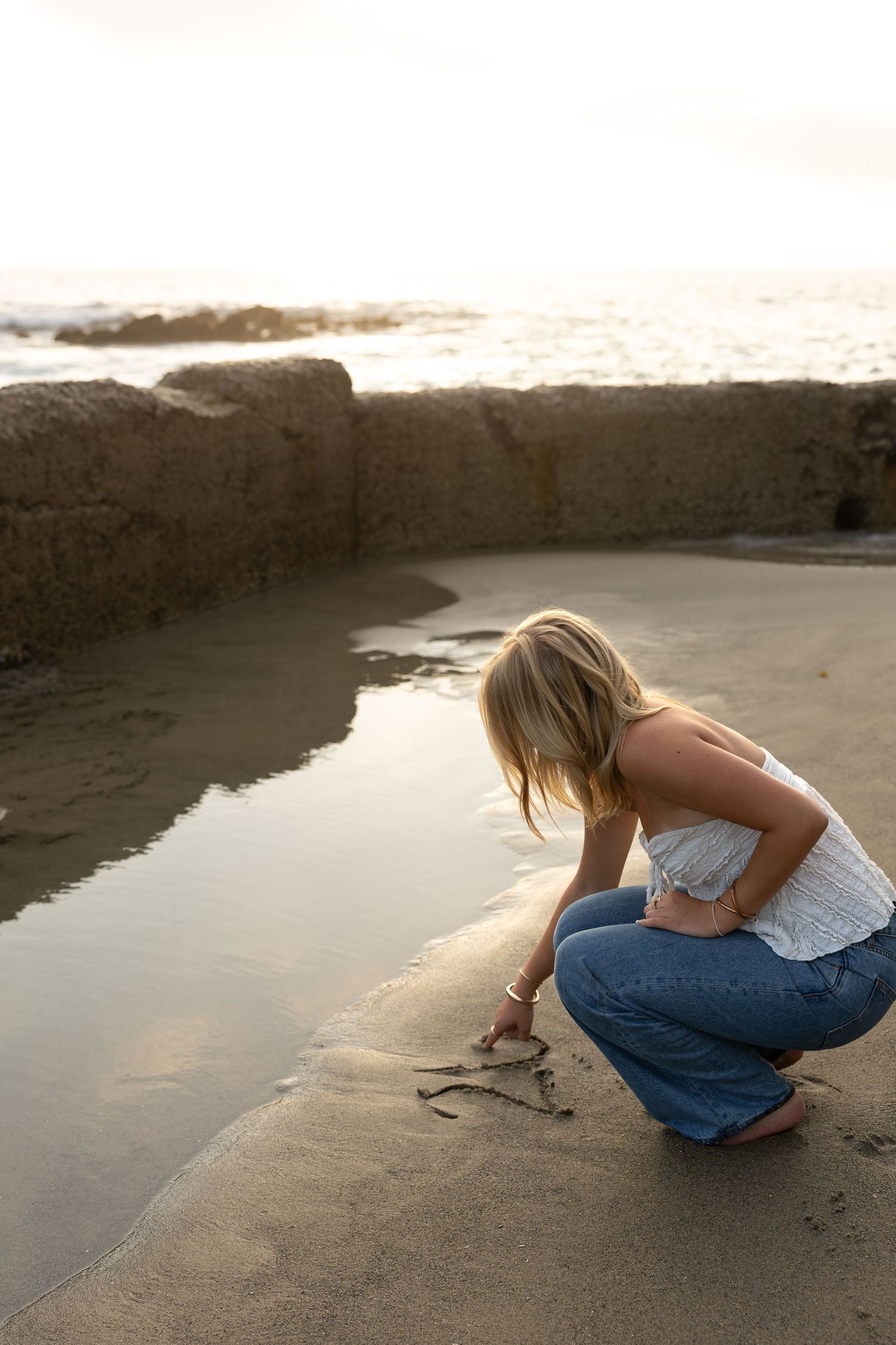 Blonde woman crouching on sandy beach at sunset, writing in the sand near a stone wall by the ocean.
