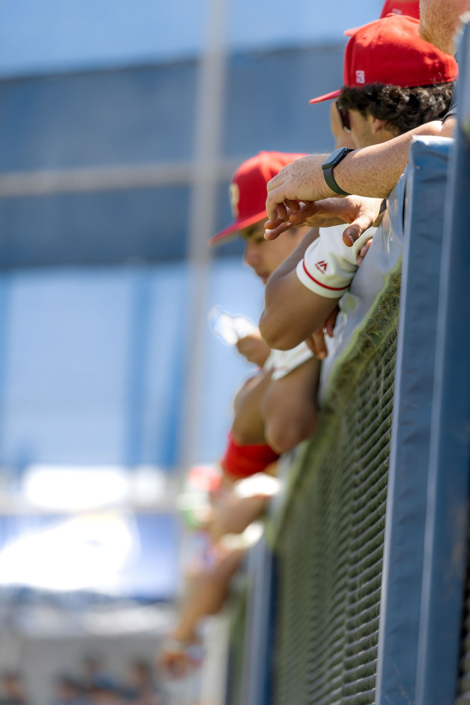Baseball players in red caps lean against dugout fence during a sunny game day.