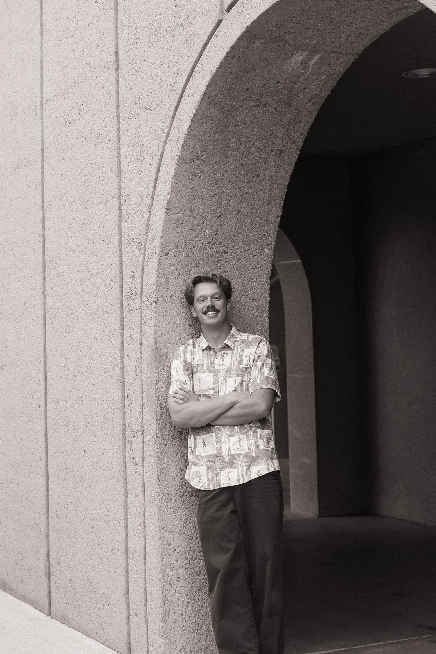 Black and white photo of a smiling man with mustache leaning against a stone arch building with arms crossed.