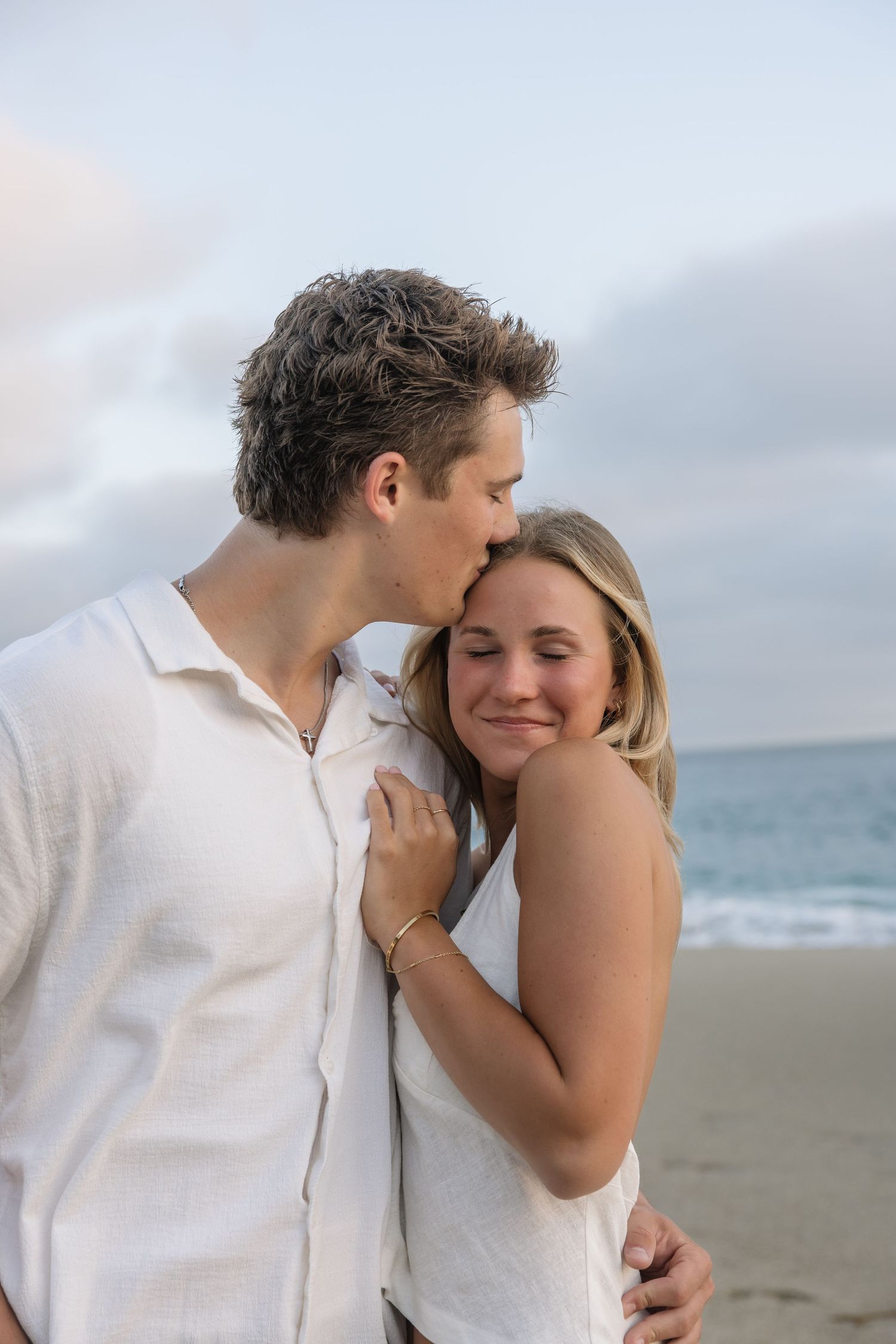 A couple shares a tender moment on the beach, with the man kissing the woman's temple as she smiles softly.