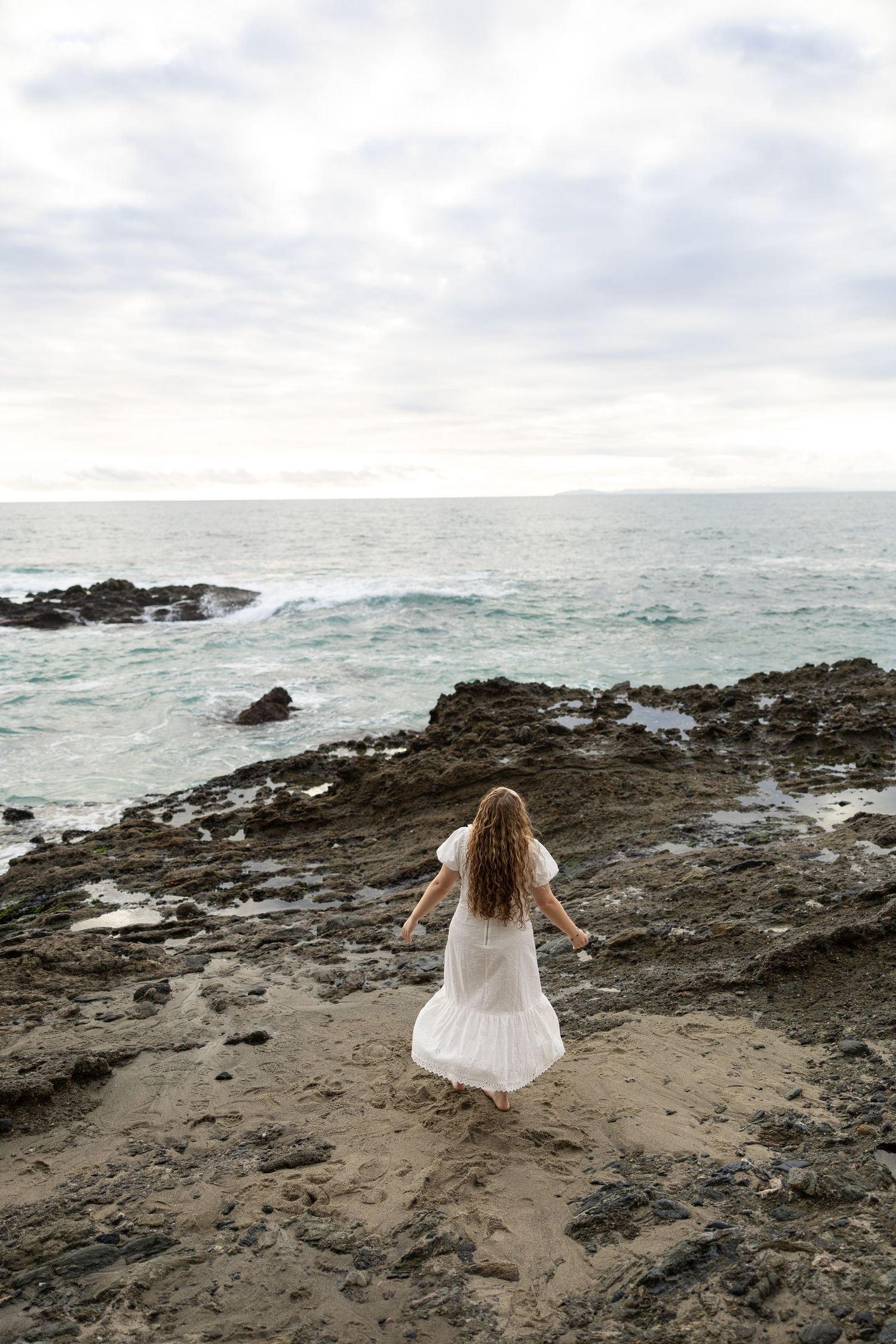 Figure in white dress explores rocky coastal beach landscape during overcast day.