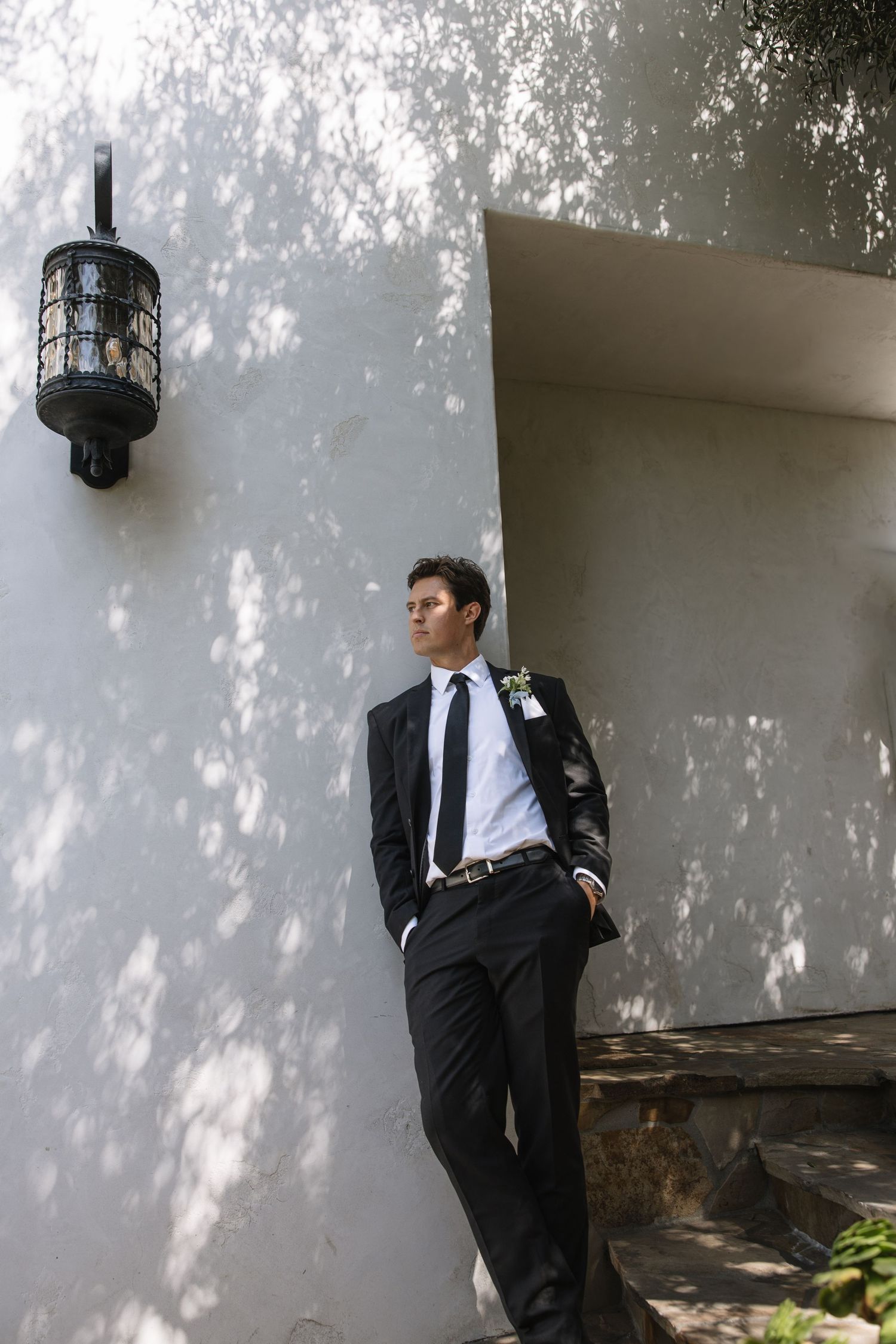Groom in black suit with boutonniere leaning against white wall outdoors, dappled sunlight creating shadows.