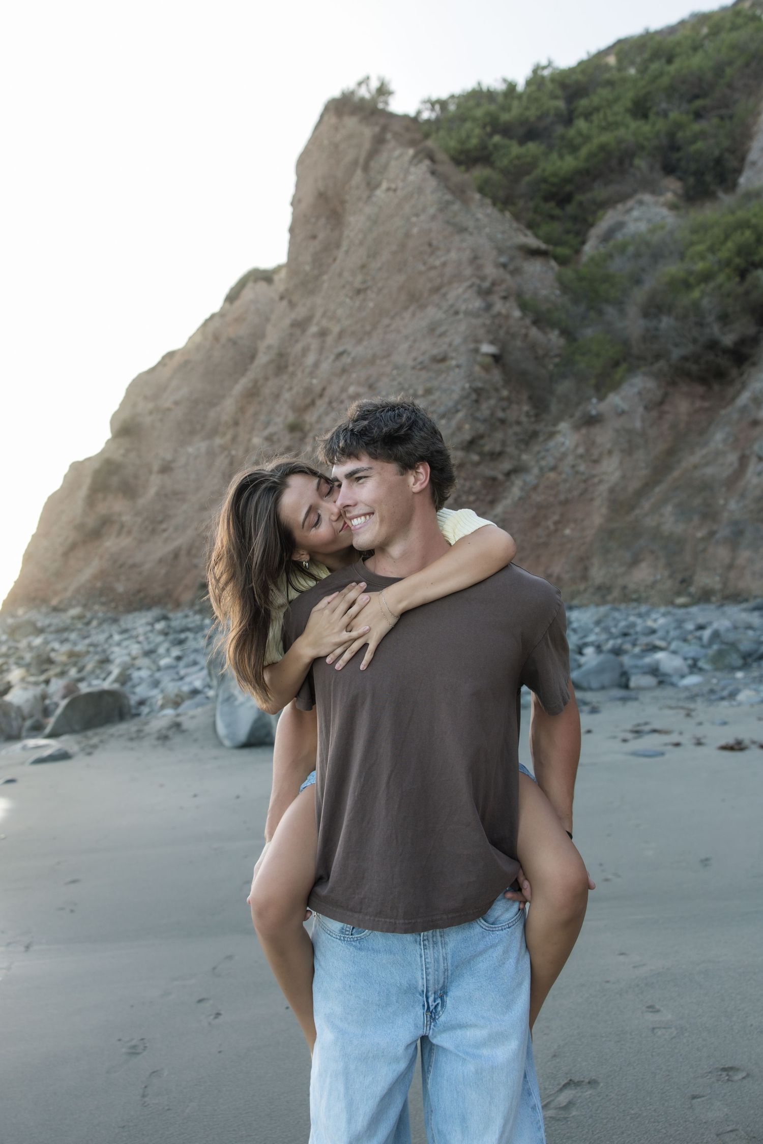 A smiling young man gives a woman a piggyback ride on a rocky beach as she kisses his cheek, with a rugged cliff behind them.