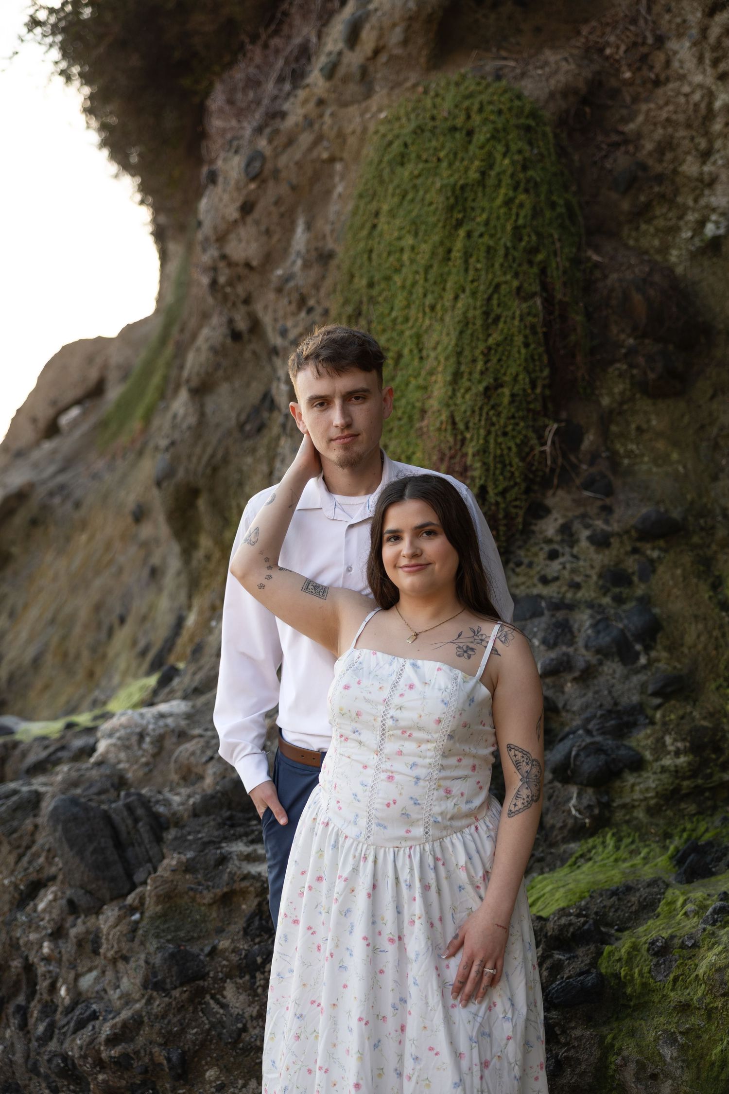 A romantic couple poses against a rocky coastal cliff backdrop during a sunset photoshoot.