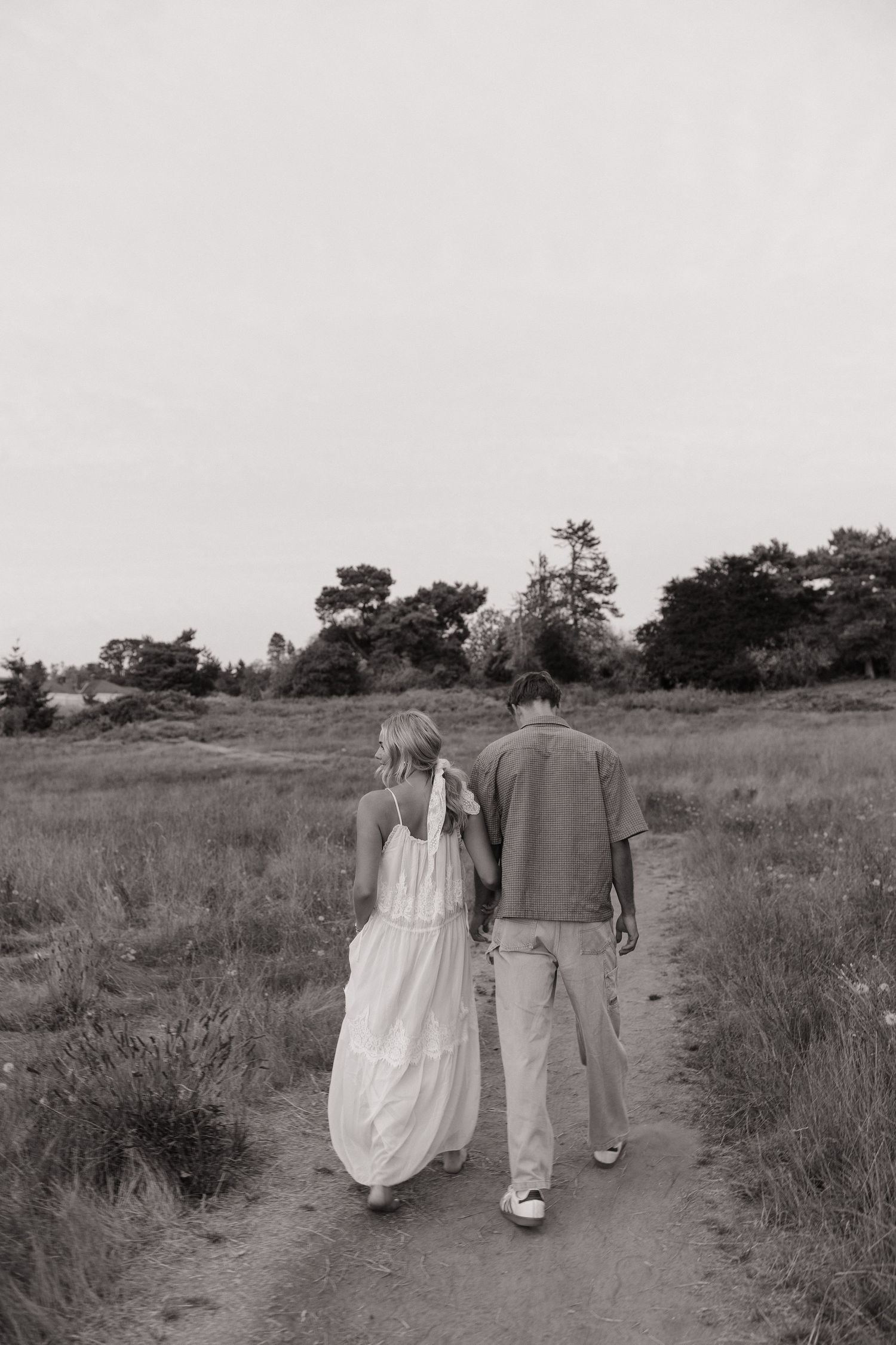 Black and white photo of a couple walking hand in hand down a dirt path through a scenic meadow with trees.