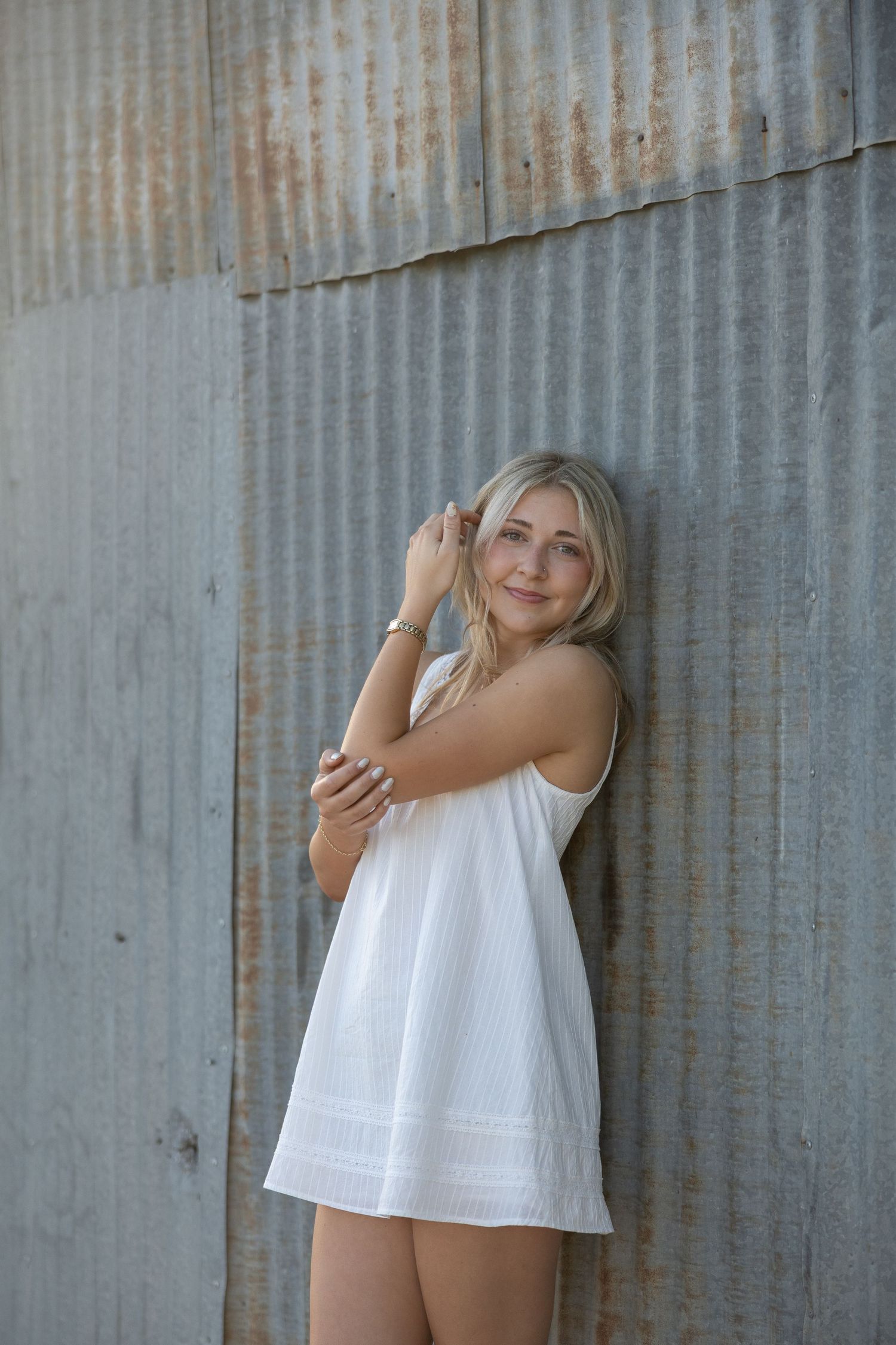 Young blonde woman in white sundress posing against rustic corrugated metal wall, smiling softly.