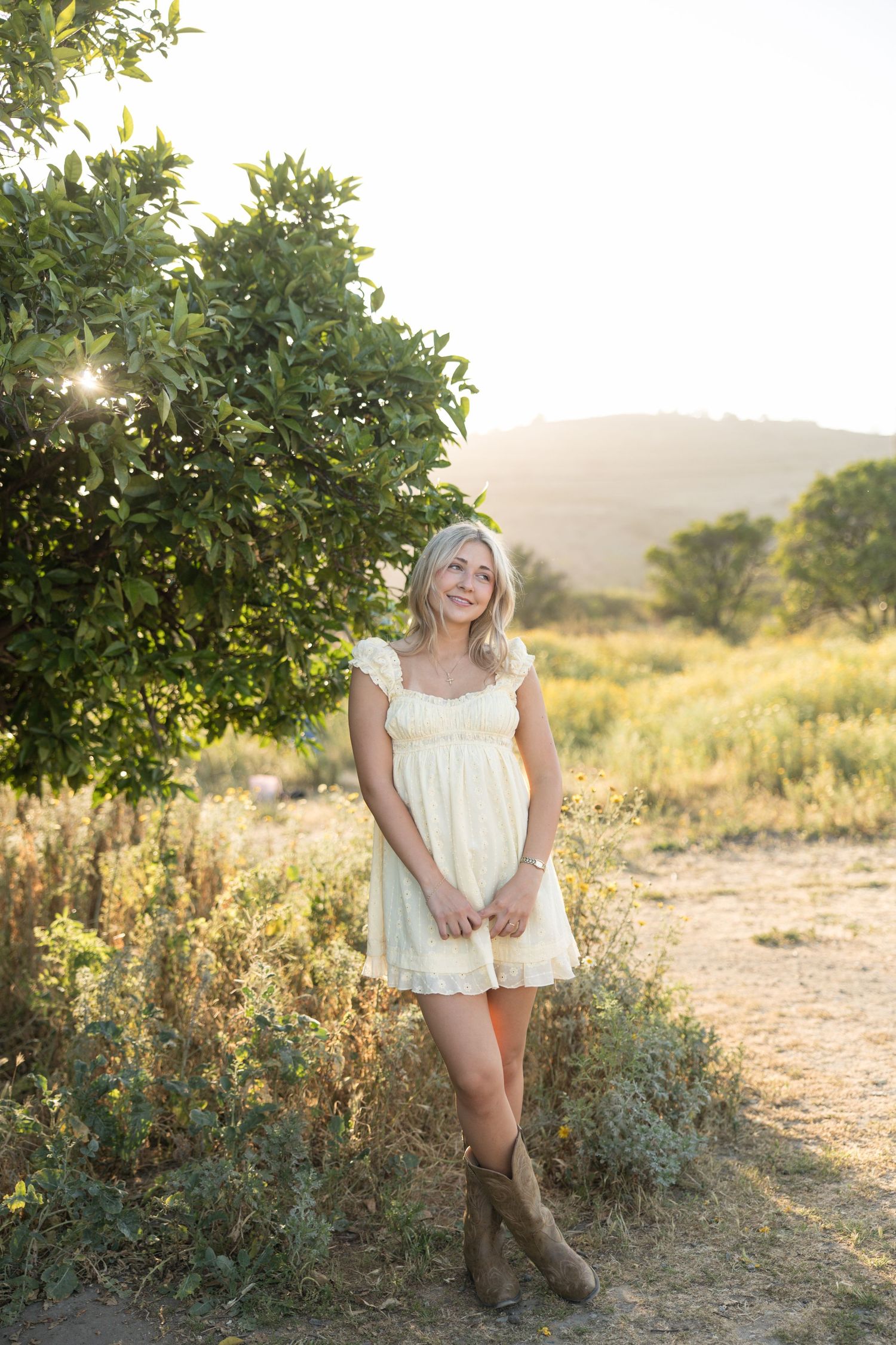 Blonde woman in white dress and brown boots standing in a sunlit field near a green tree with hills in the background.