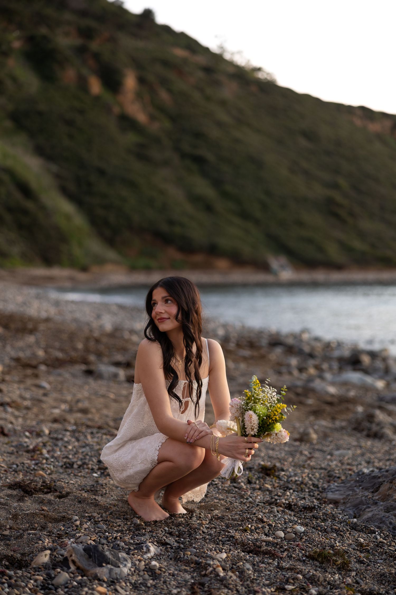 A person in lace dress crouches on a rocky beach holding wildflowers at sunset.