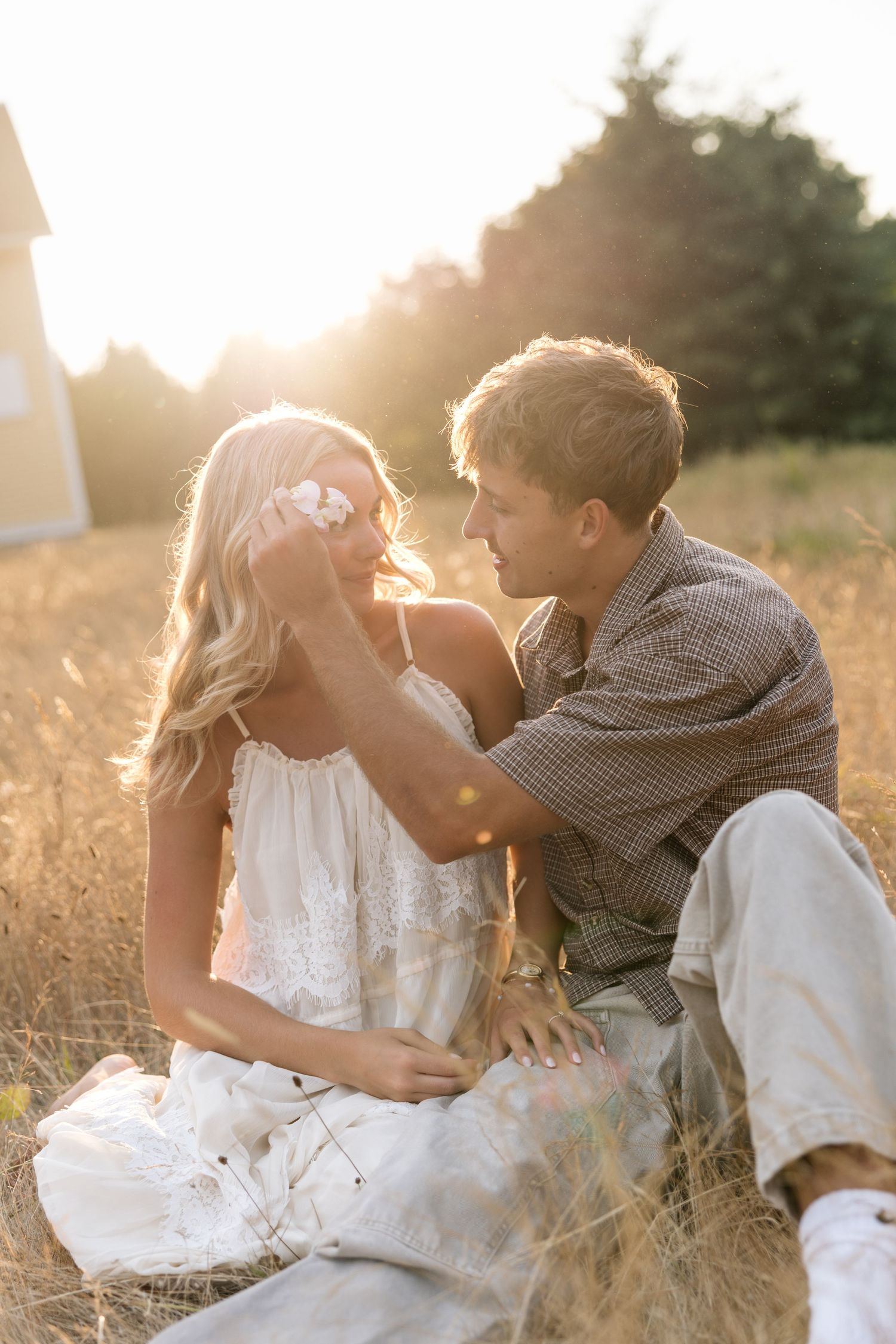 A couple sits in a golden sunlit field, the man gently tucking hair behind the woman's ear in a romantic moment.